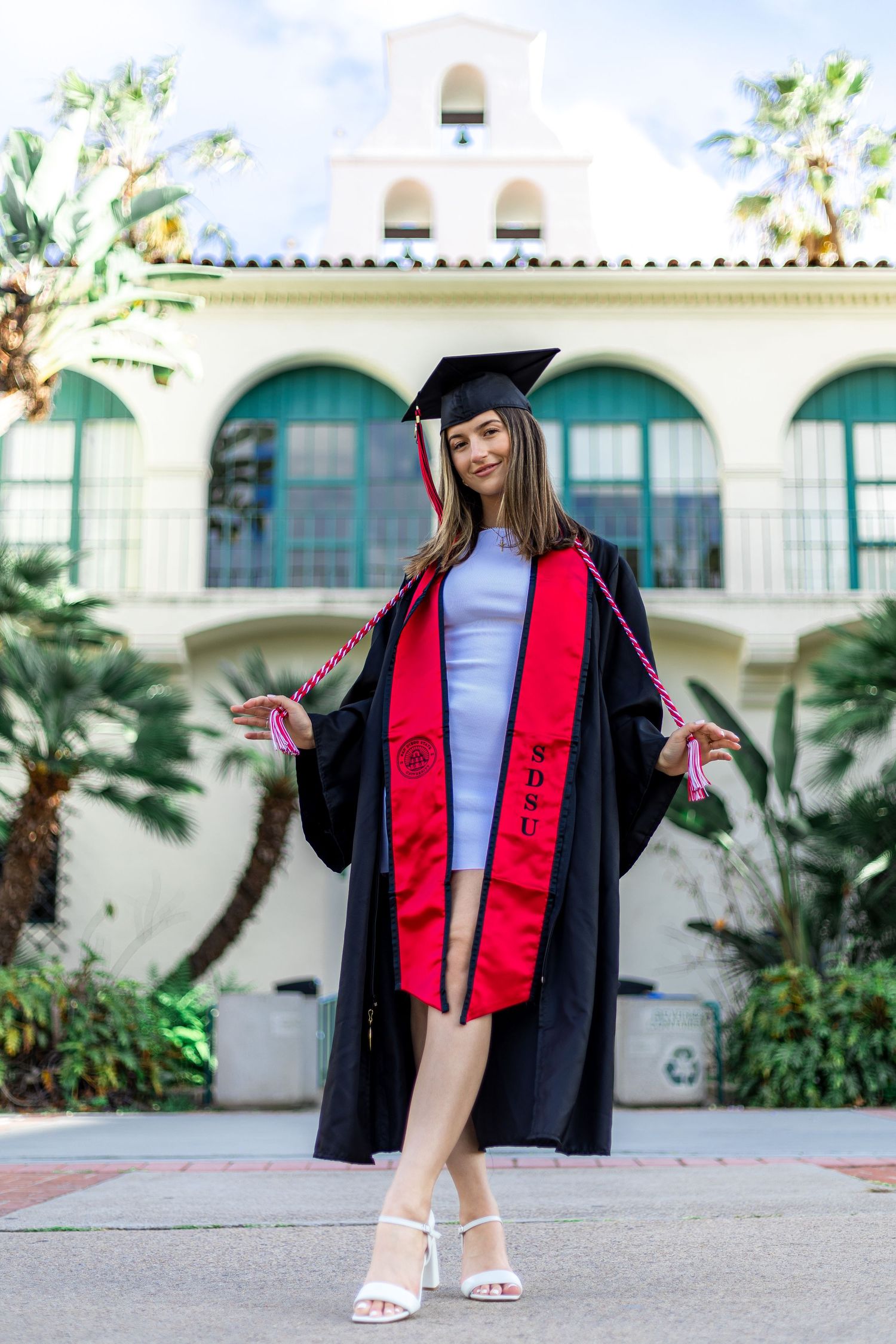Graduate in full regalia posing in front of Spanish-style campus architecture.