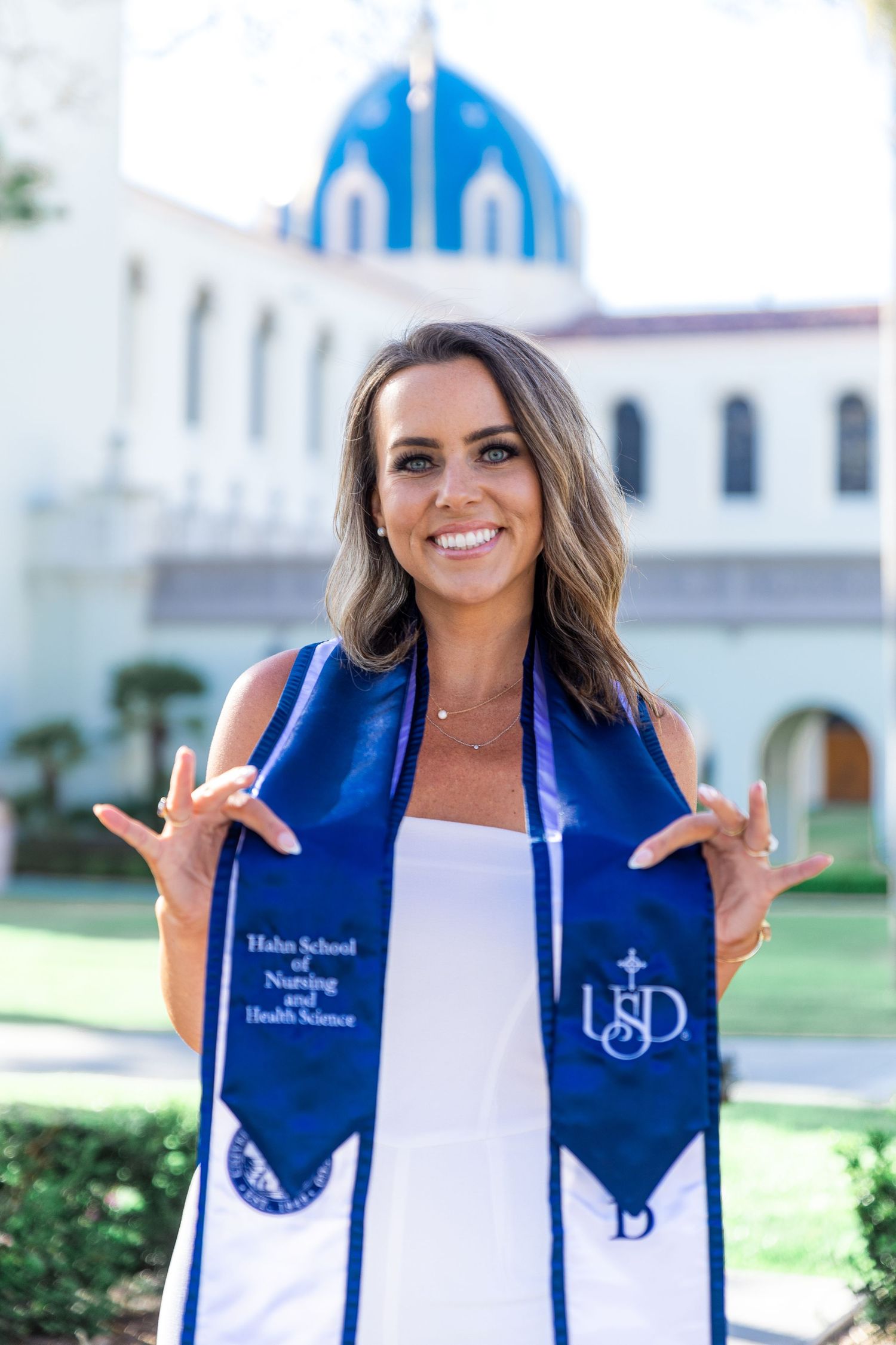 Graduate wearing blue graduation stole makes hand gestures in front of white church building with blue dome.
