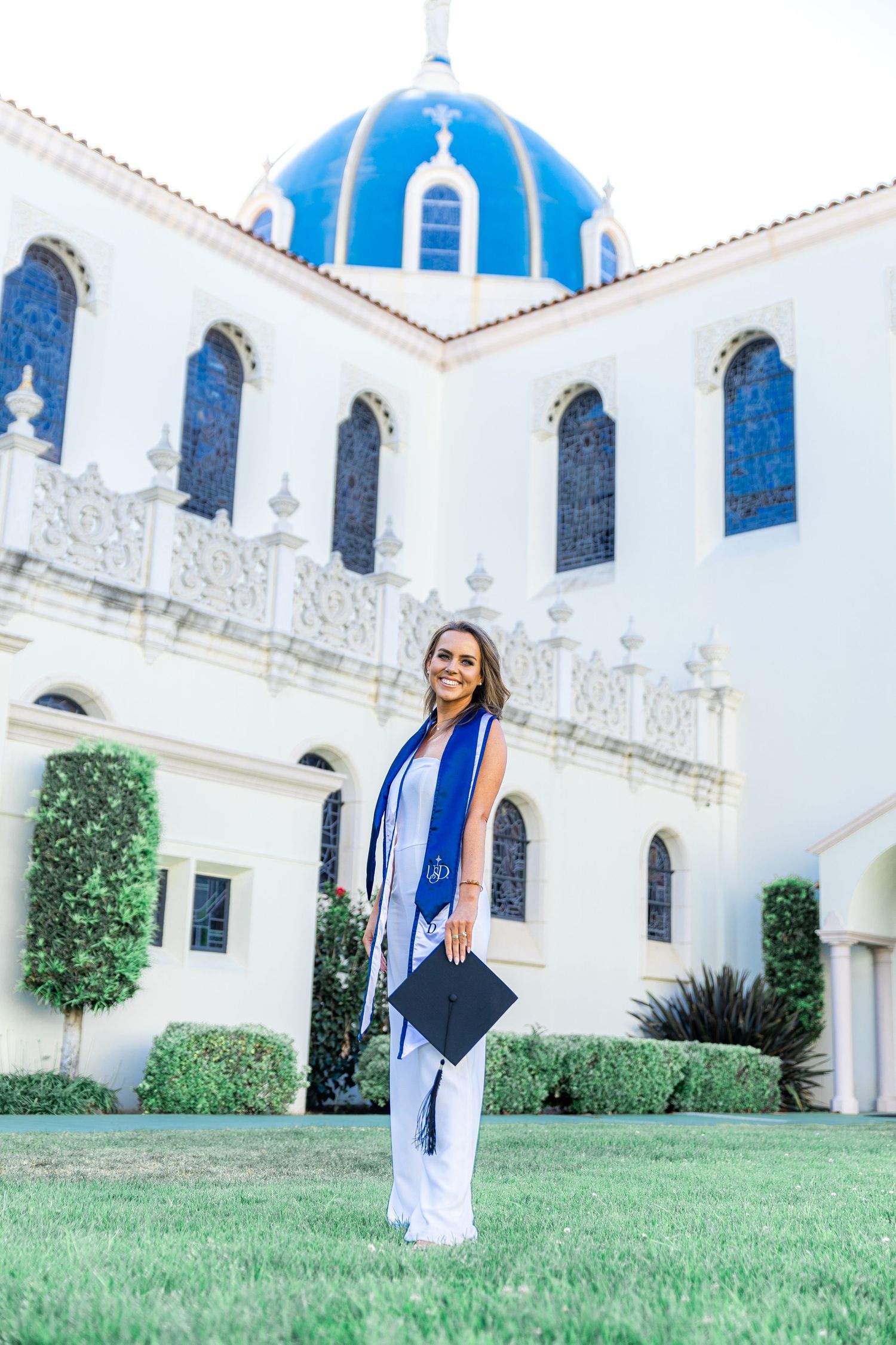 Graduate in blue and white stole poses with diploma in front of Mediterranean-style building with blue dome.