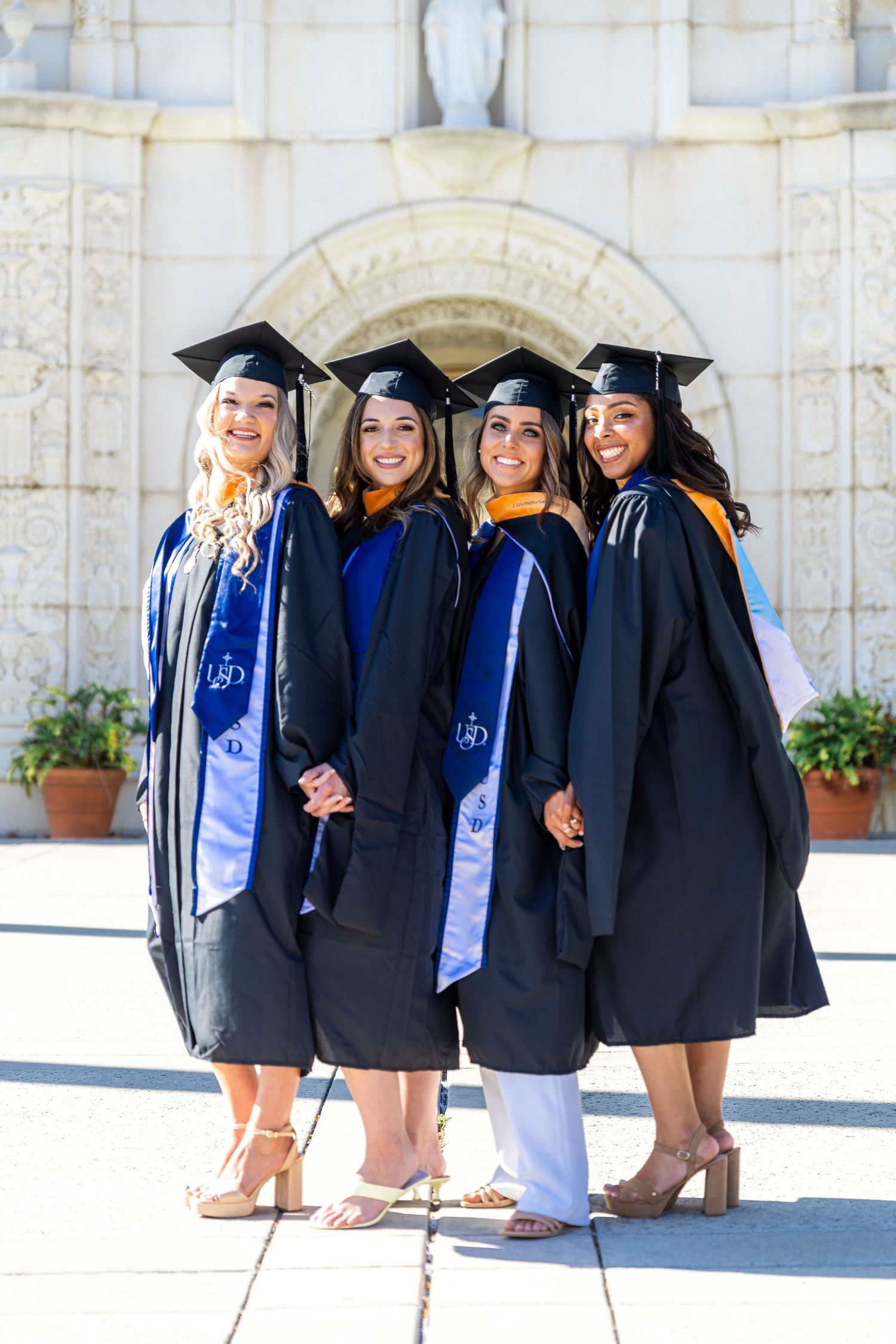 Graduates in black caps and gowns pose together in front of white mission-style architecture.