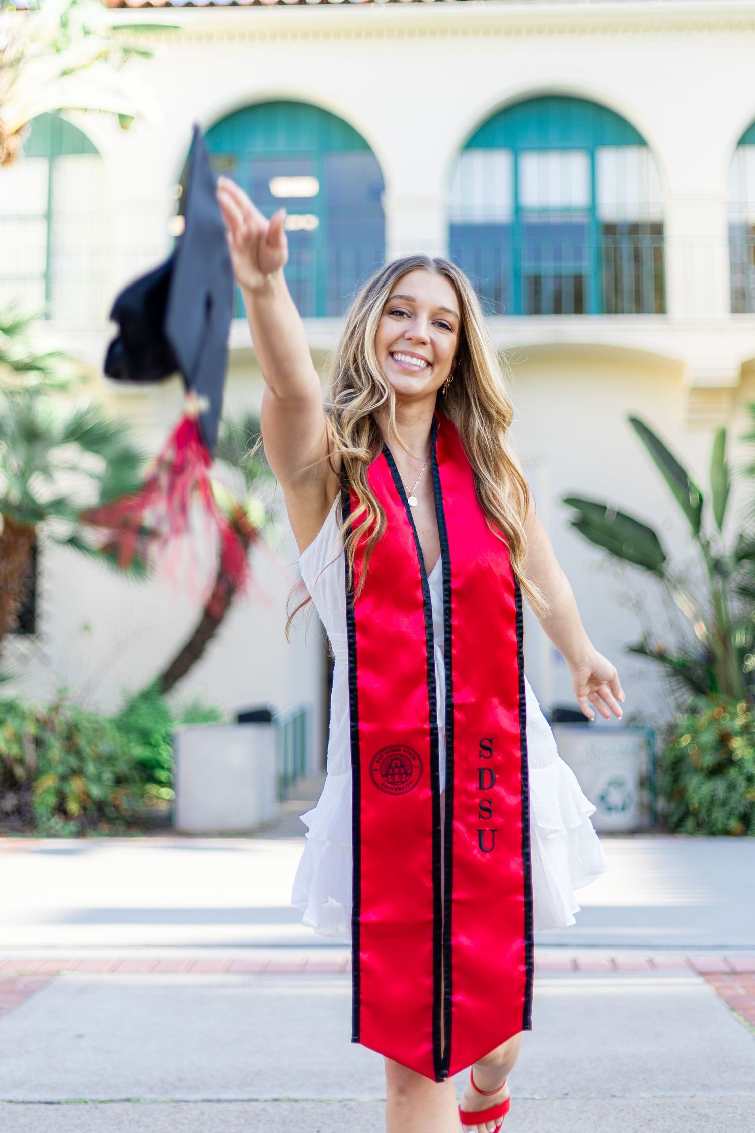 Graduate wearing a red and black graduation stole poses joyfully in an outdoor setting.