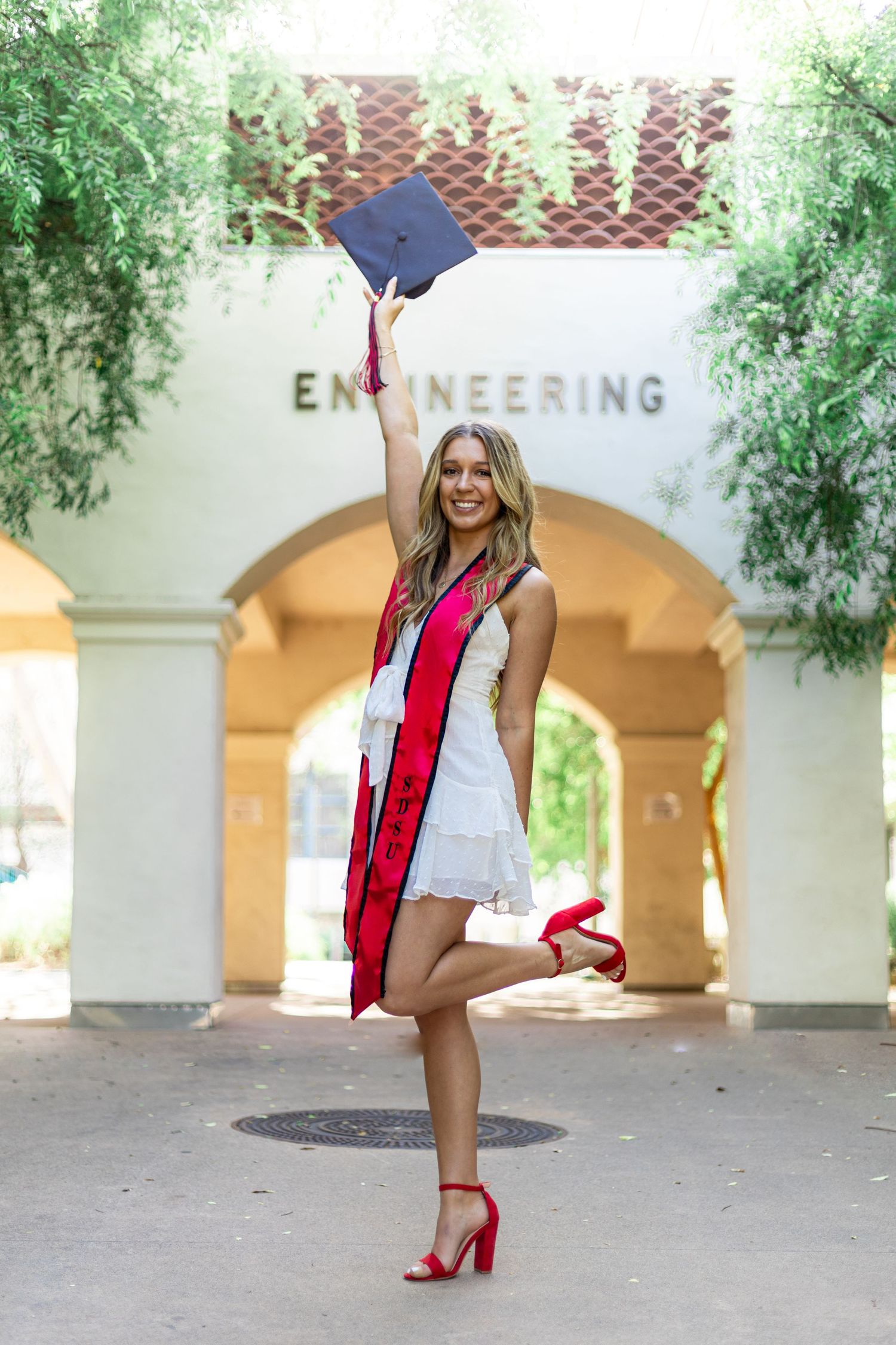 Graduate poses near Engineering building entrance in graduation attire with red heels.