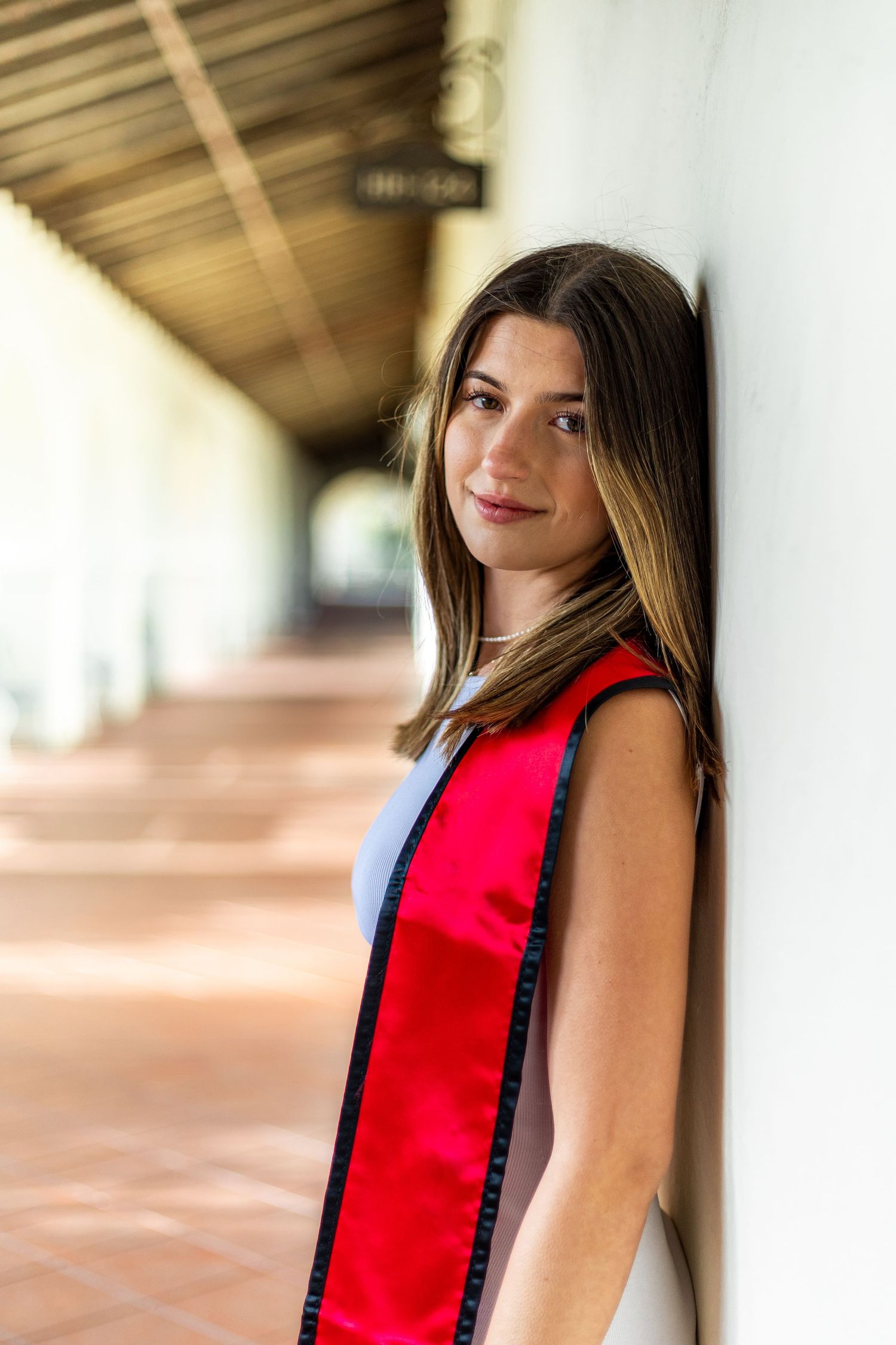 Graduate wearing red stole stands against white columned walkway in celebratory pose.