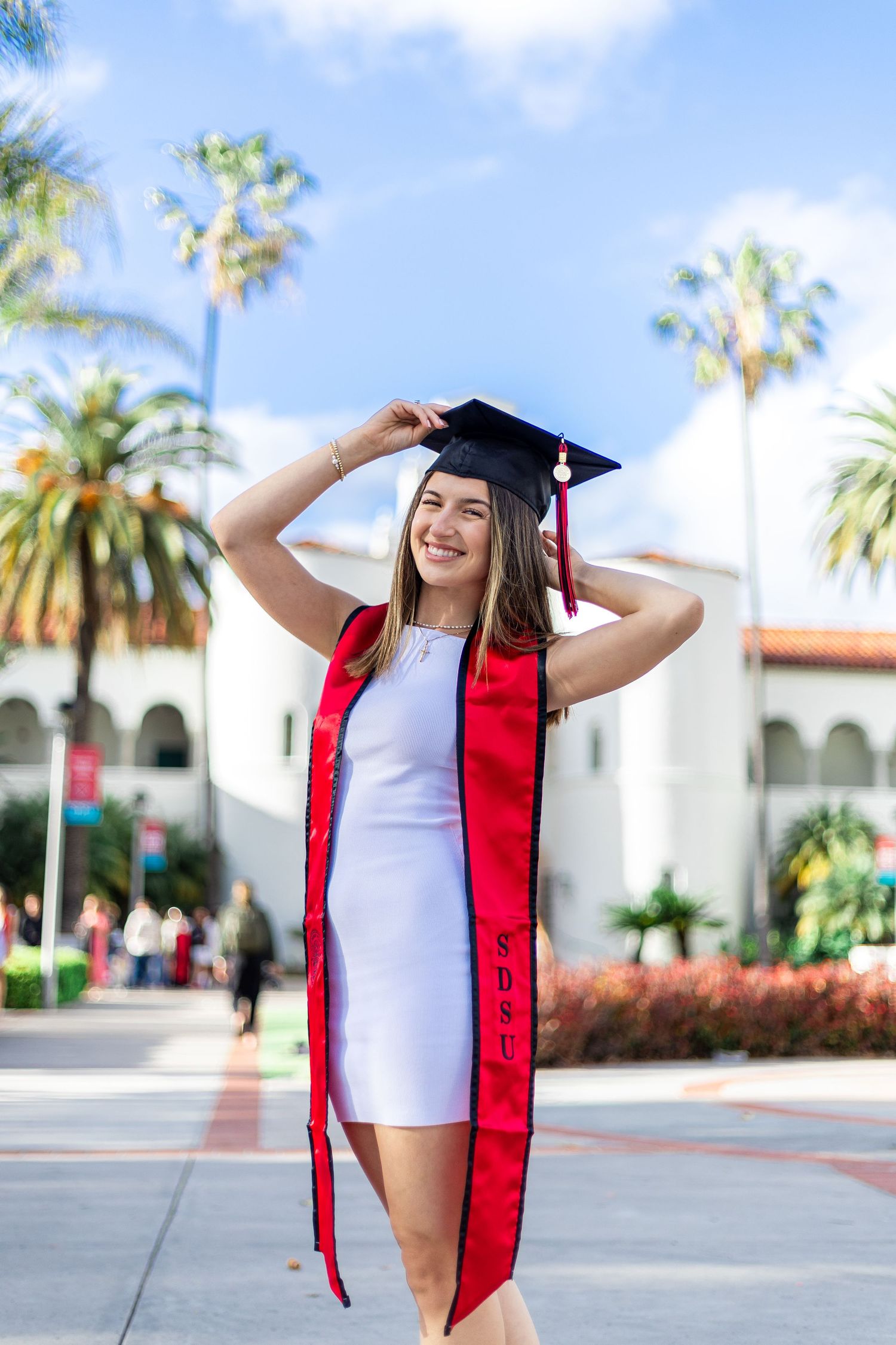 Graduate in white dress and red stole posing with cap against palm tree backdrop.