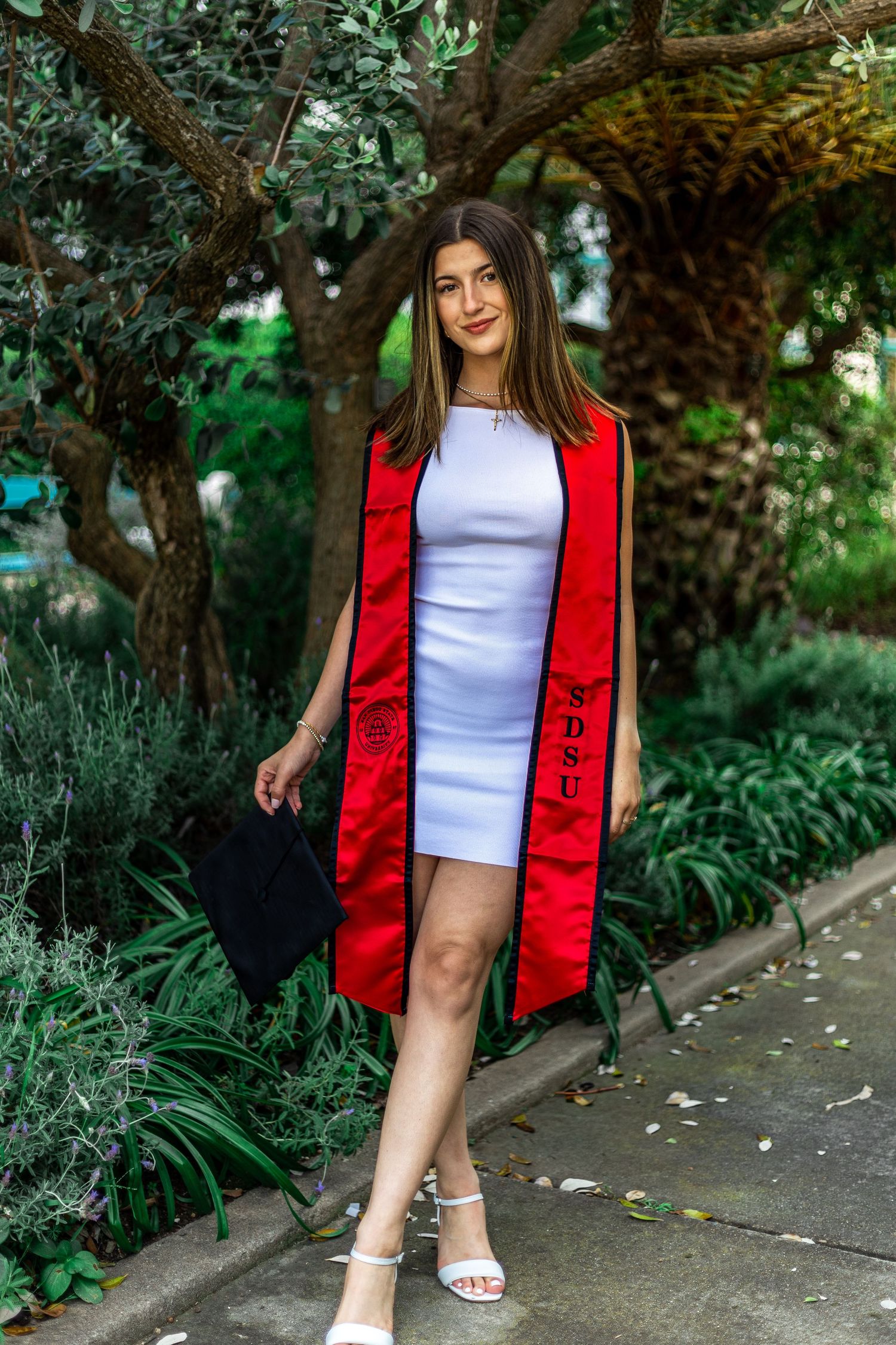 Graduate posing in white dress and red graduation stole against garden background.