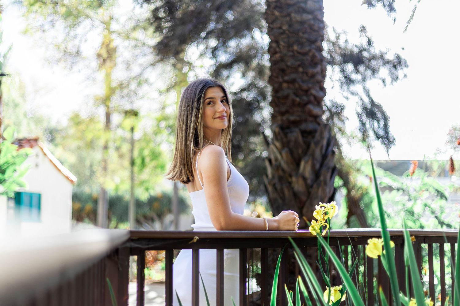 Graduate in white dress standing on outdoor balcony with scenic background.
