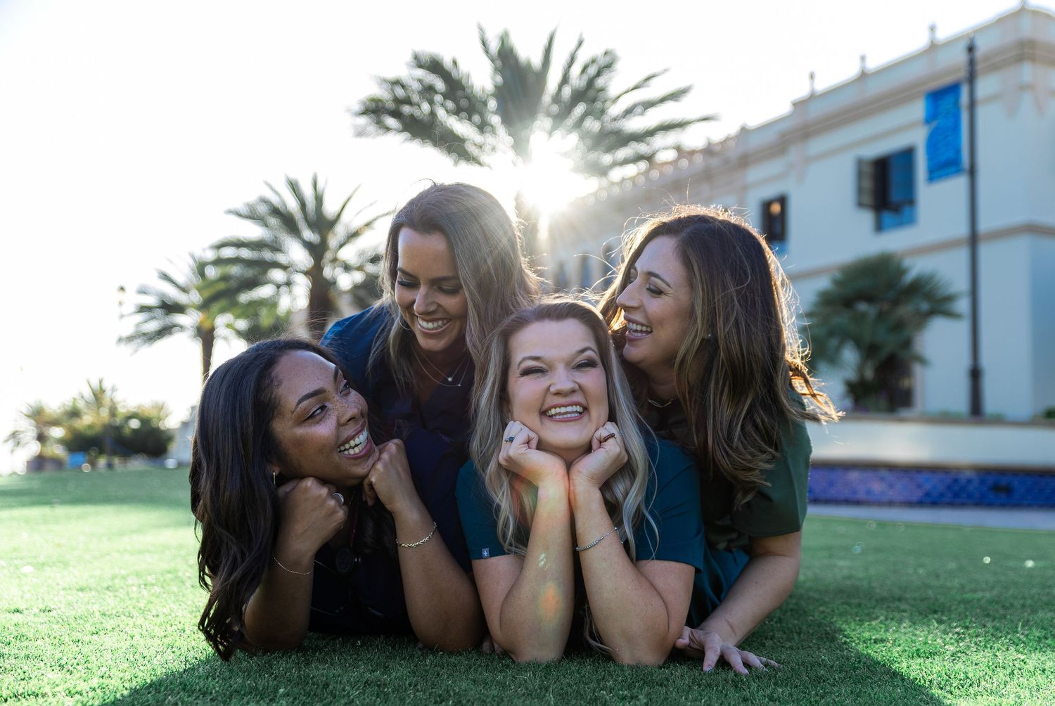 Friends in graduation attire lay on grass laughing together during sunset photo session.