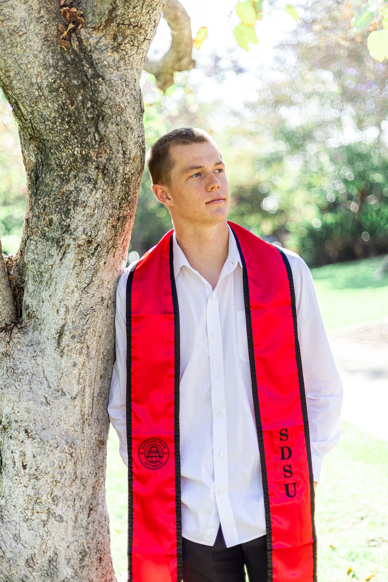 Graduate in white shirt and red stole poses against tree backdrop.