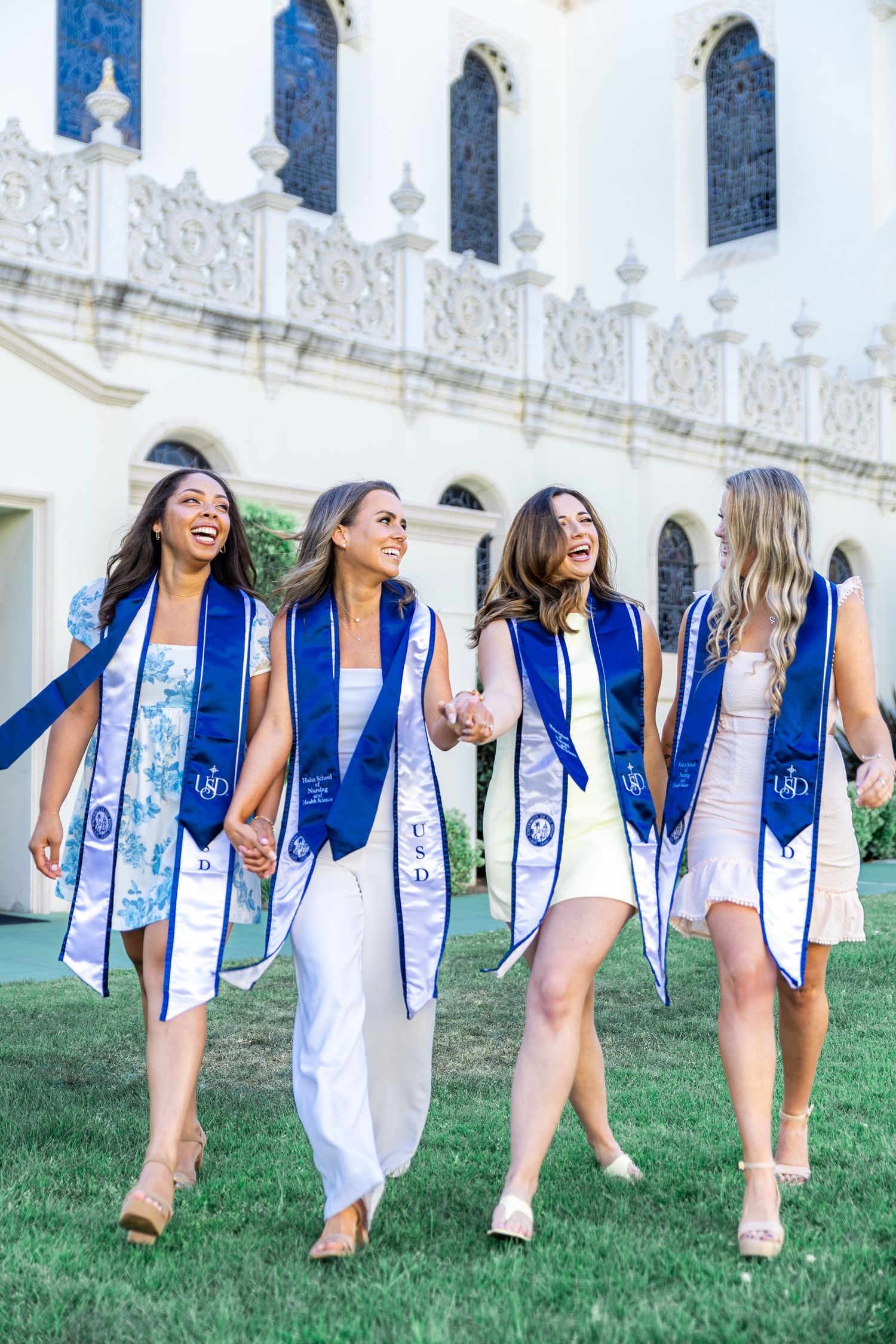 Graduates in white dresses and blue stoles laugh while walking together on campus lawn.