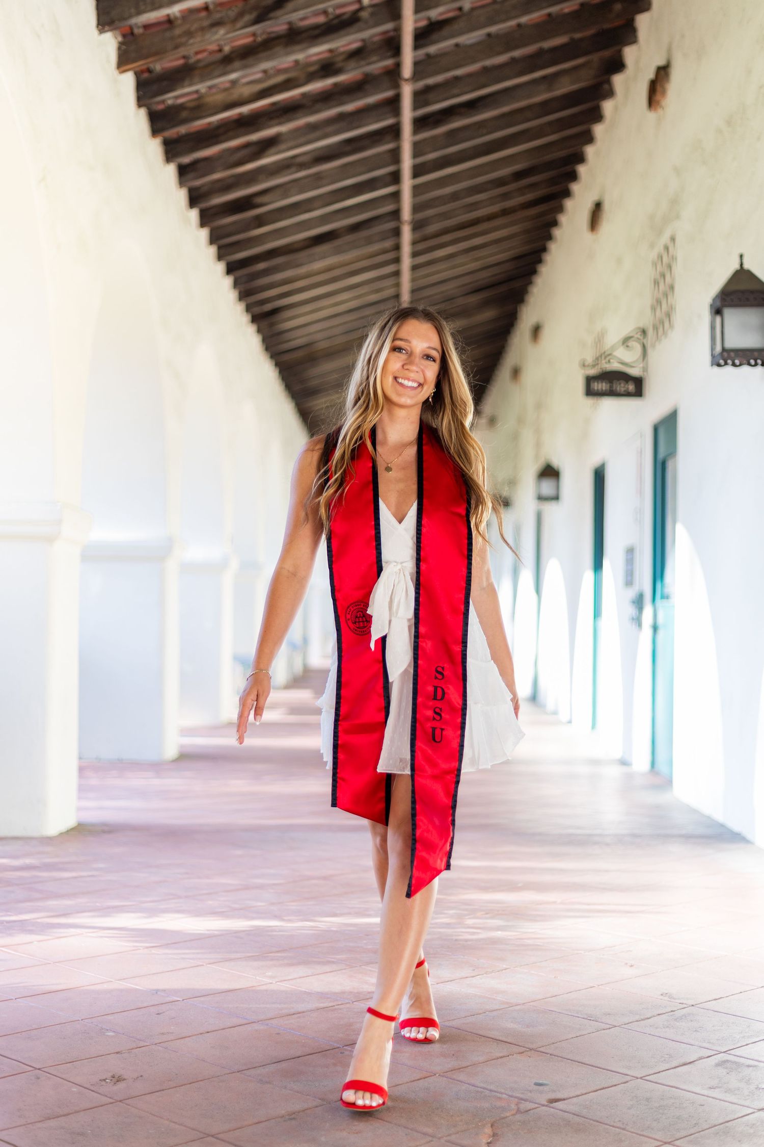 Person in red graduation stole twirls playfully in white columned hallway.