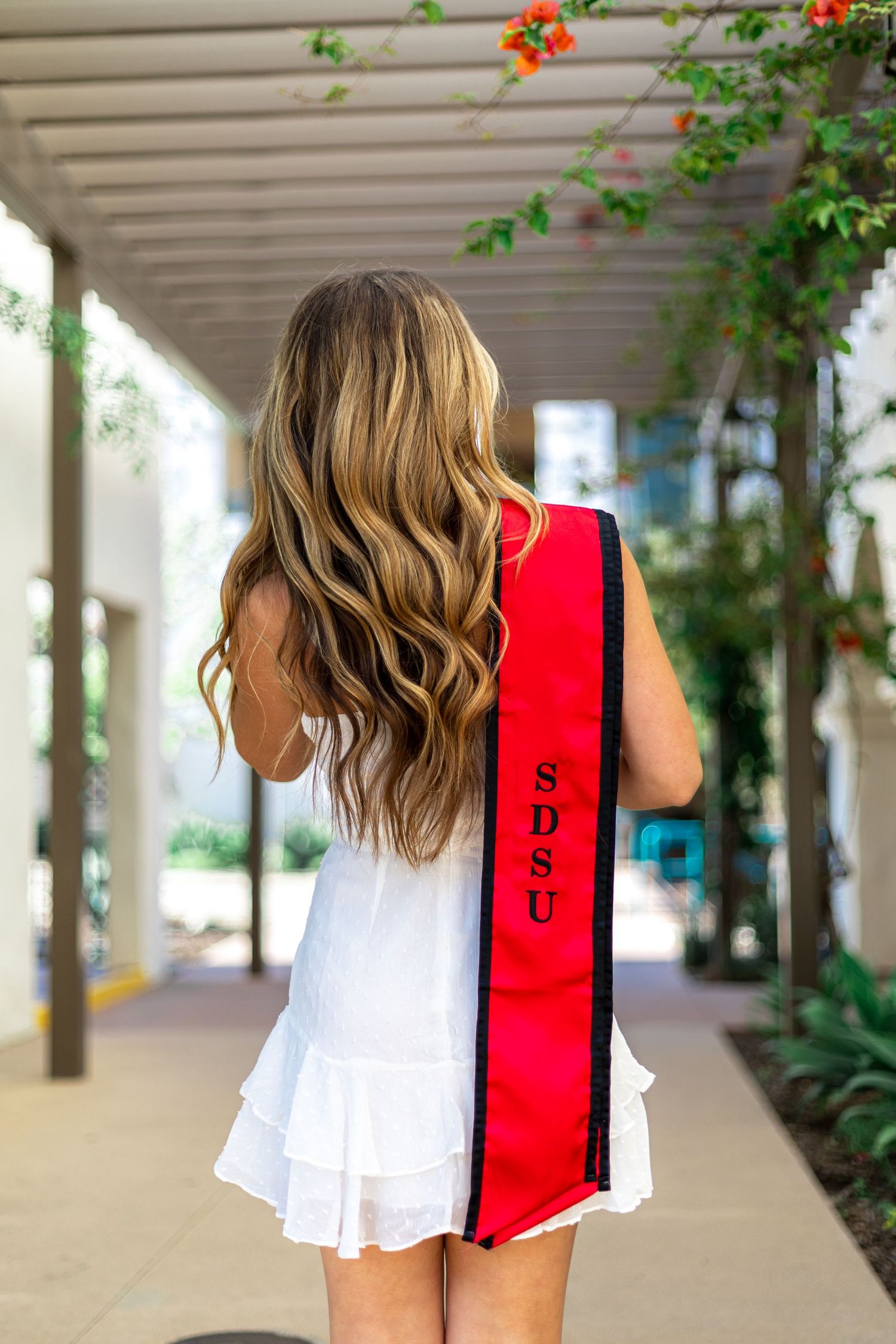 Graduate in white dress and red stole walks through outdoor colonnade with flowering vines.