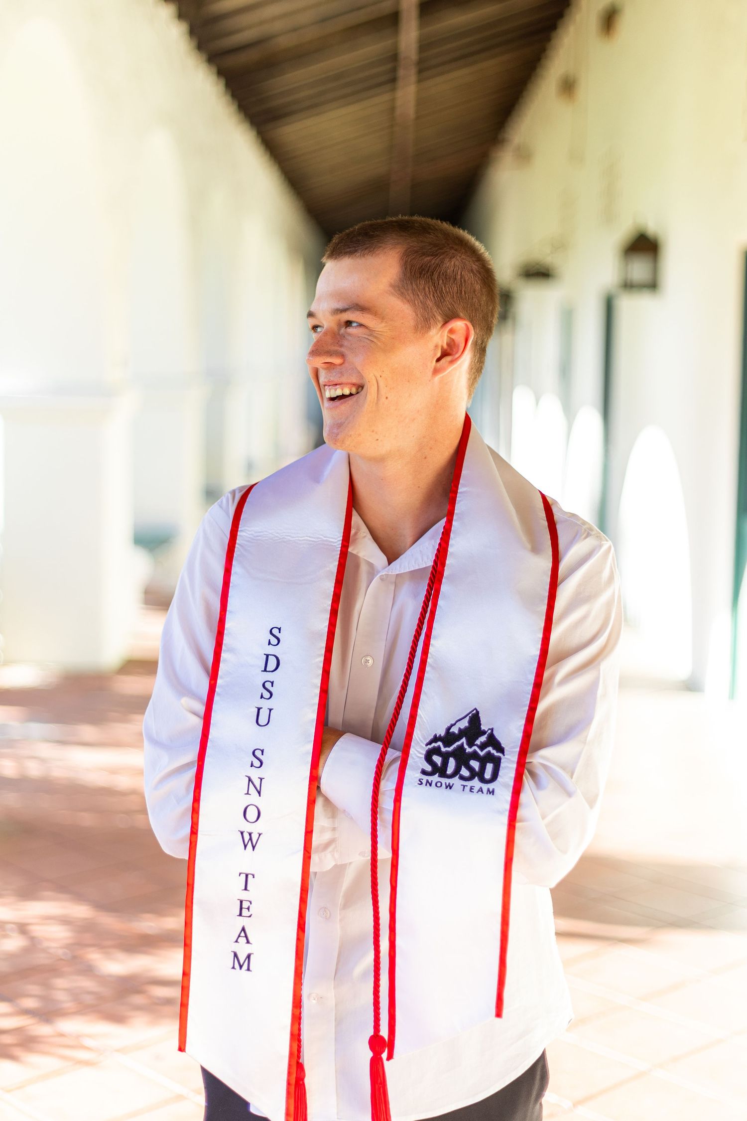Graduate in white shirt with embroidered stole poses in covered walkway.