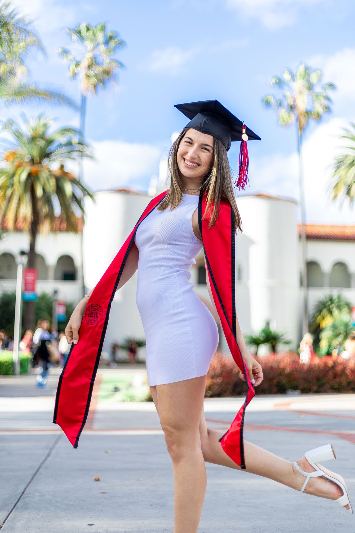 Graduate in cap and gown twirling on campus with palm trees in background.
