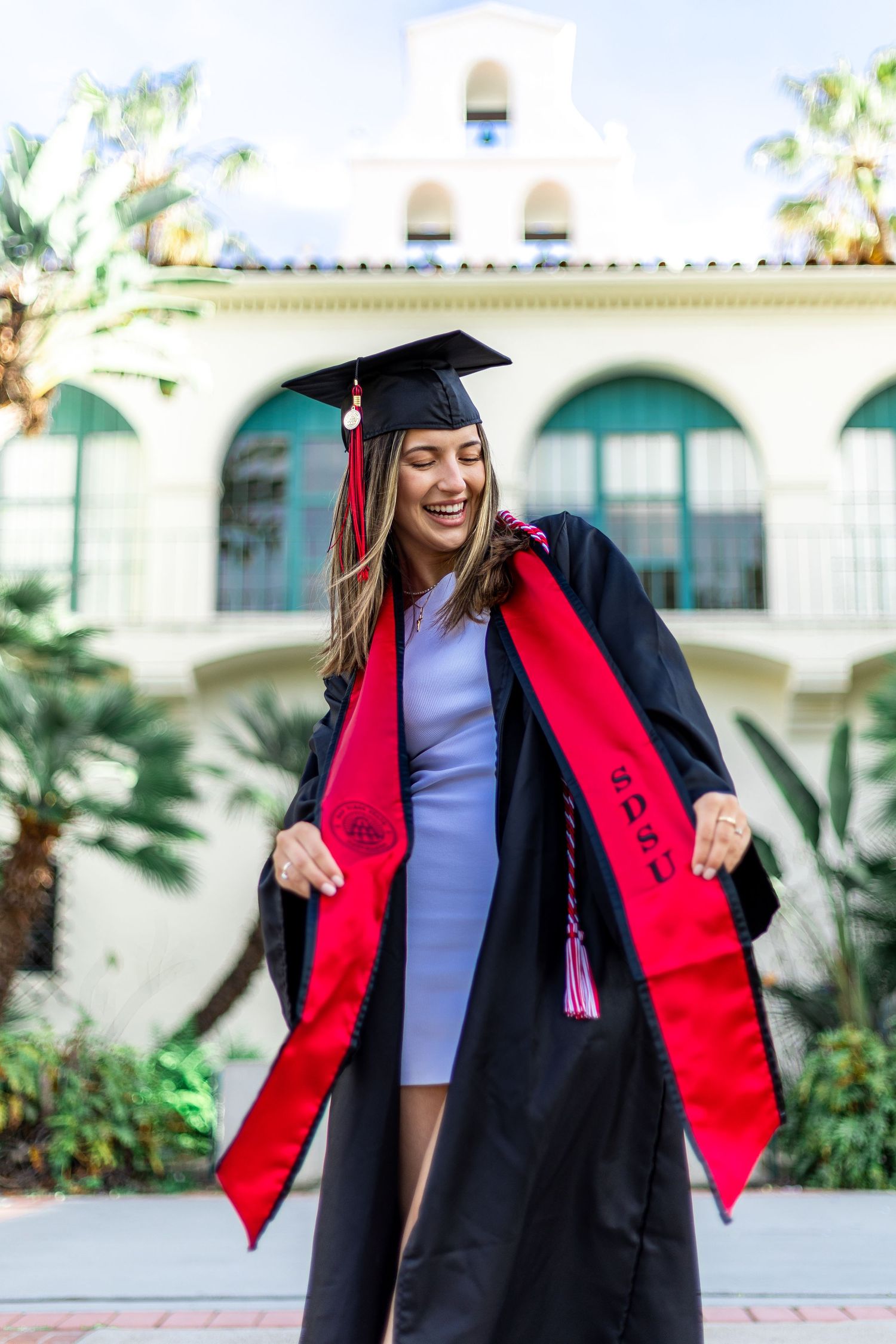 Graduate in cap and gown with red stole celebrates near Spanish-style architecture.