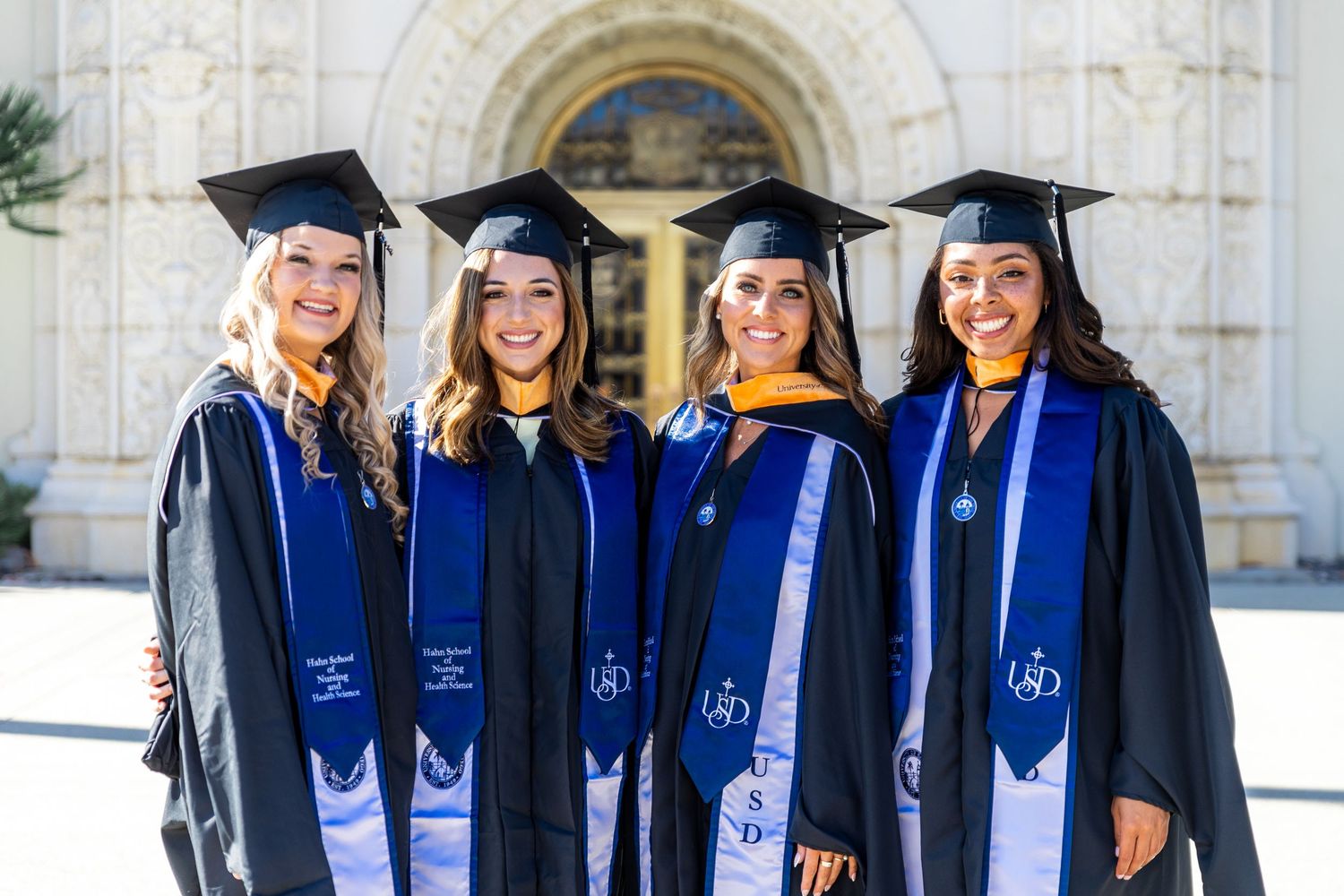 Graduates in academic regalia pose together in front of ornate white building during commencement.