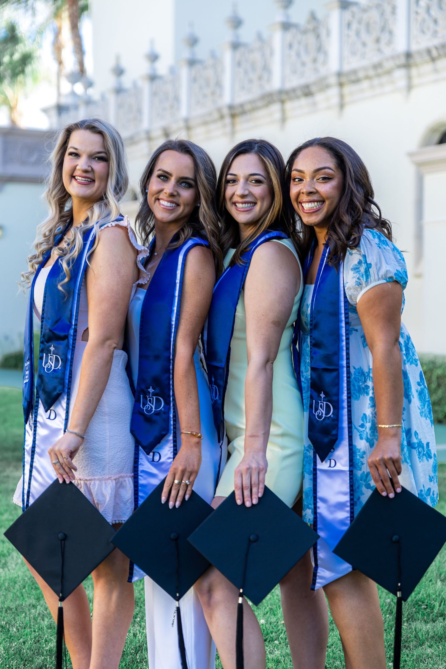 Four graduates in blue stoles and black caps pose together holding their graduation caps on a sunny day.