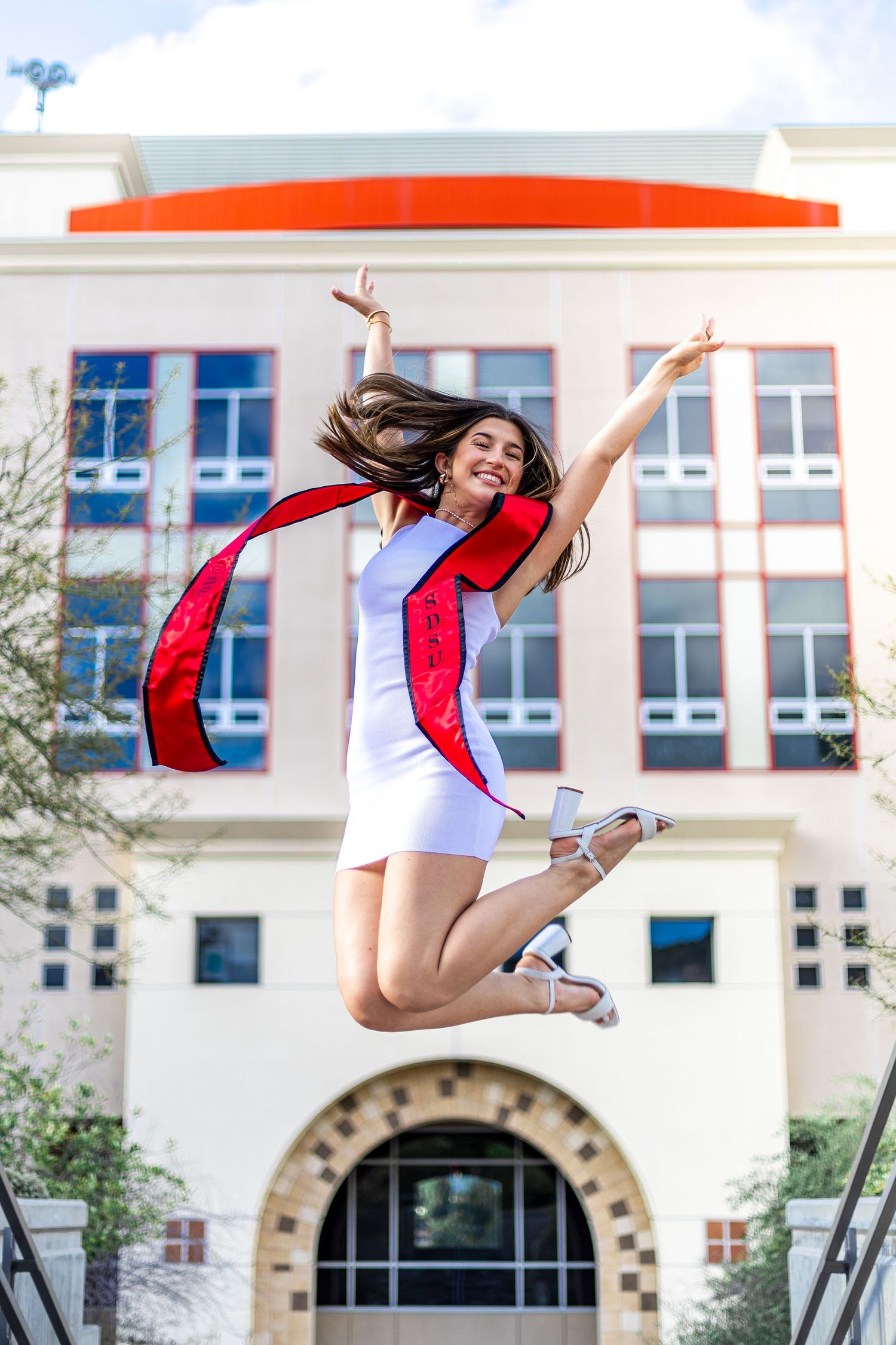 Excited graduate jumping high in red stole and white dress in front of campus building.