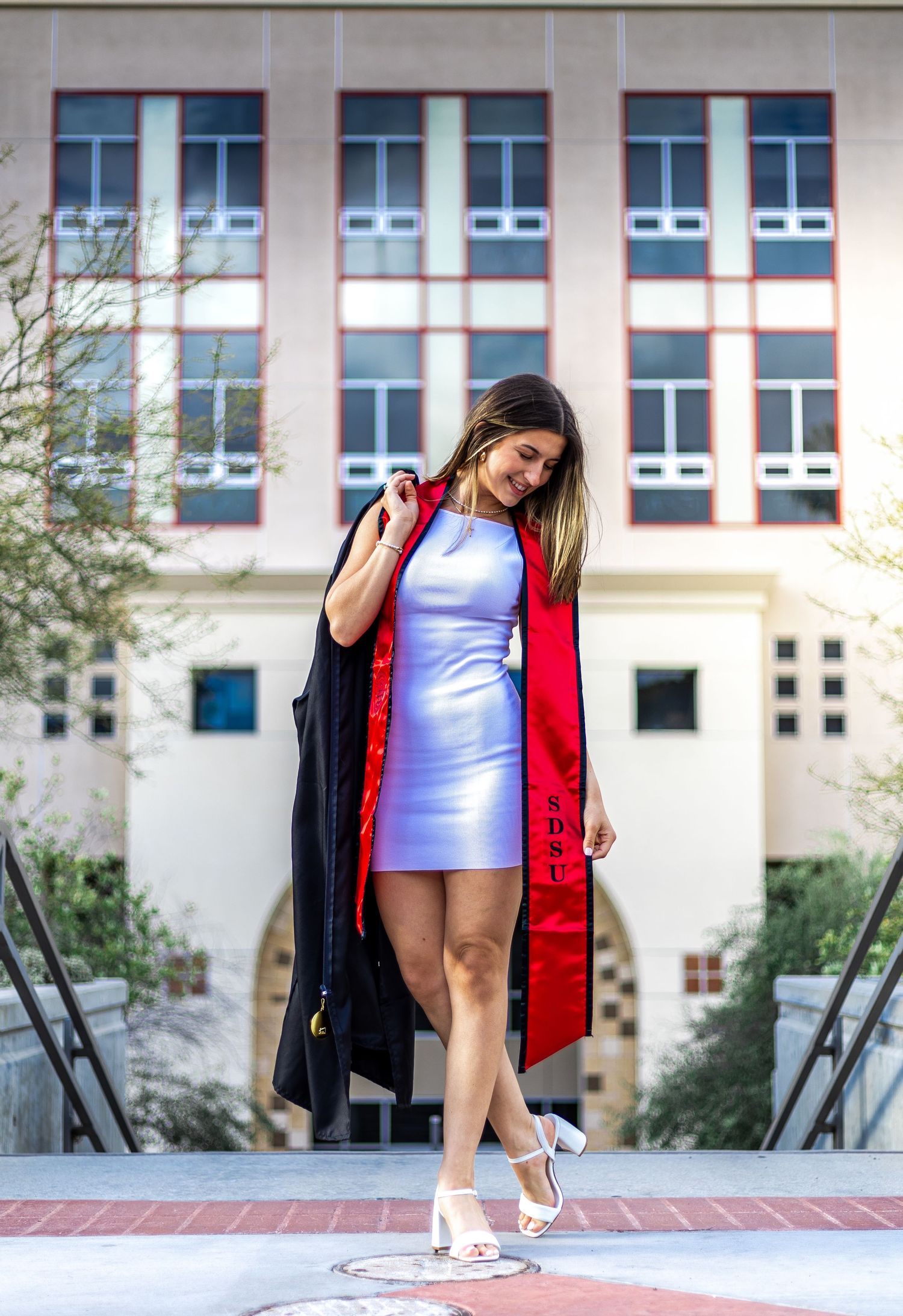 Graduate in black gown and red stole poses against modern building exterior.