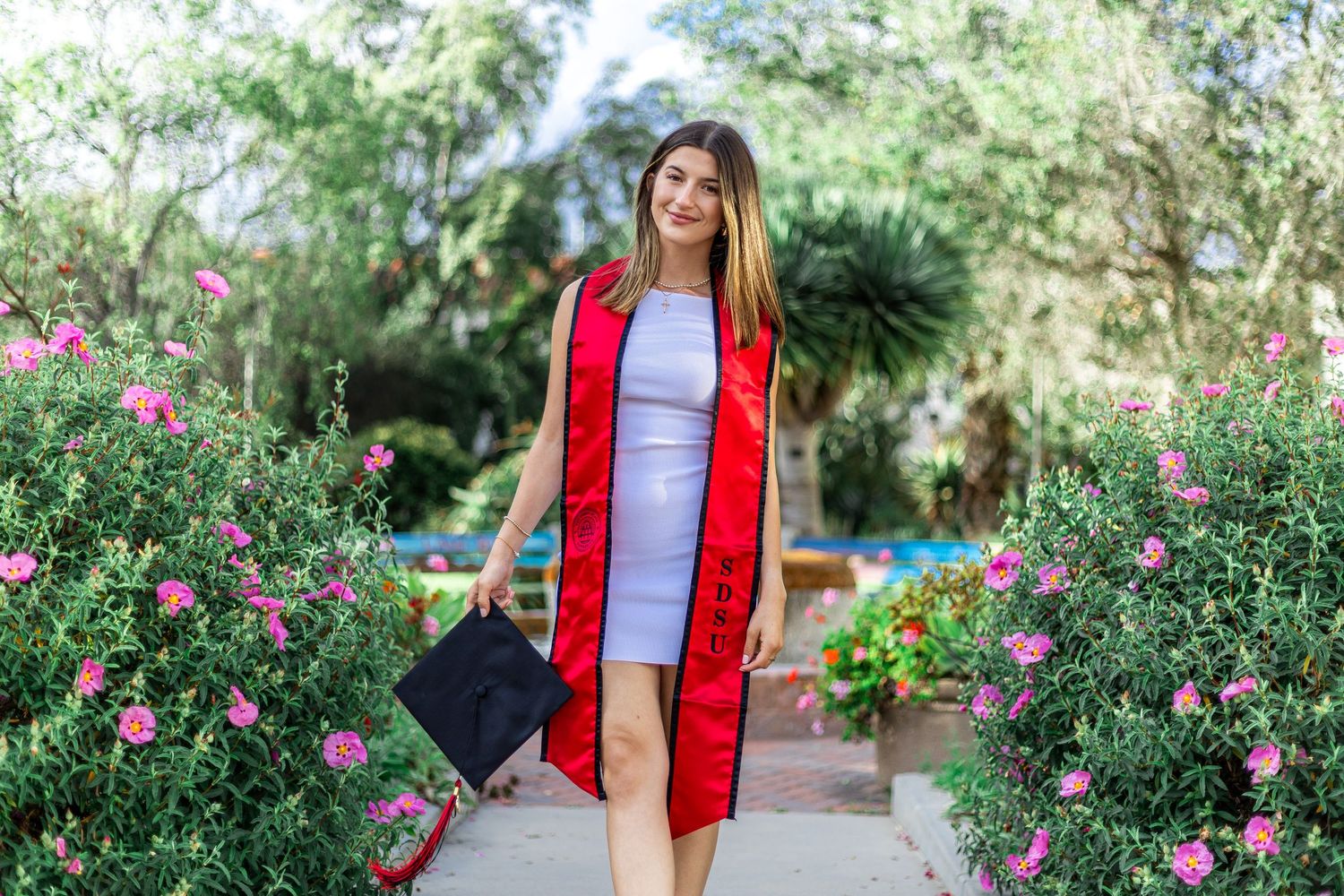 Graduate in red stole holding cap while walking through garden with pink flowers.