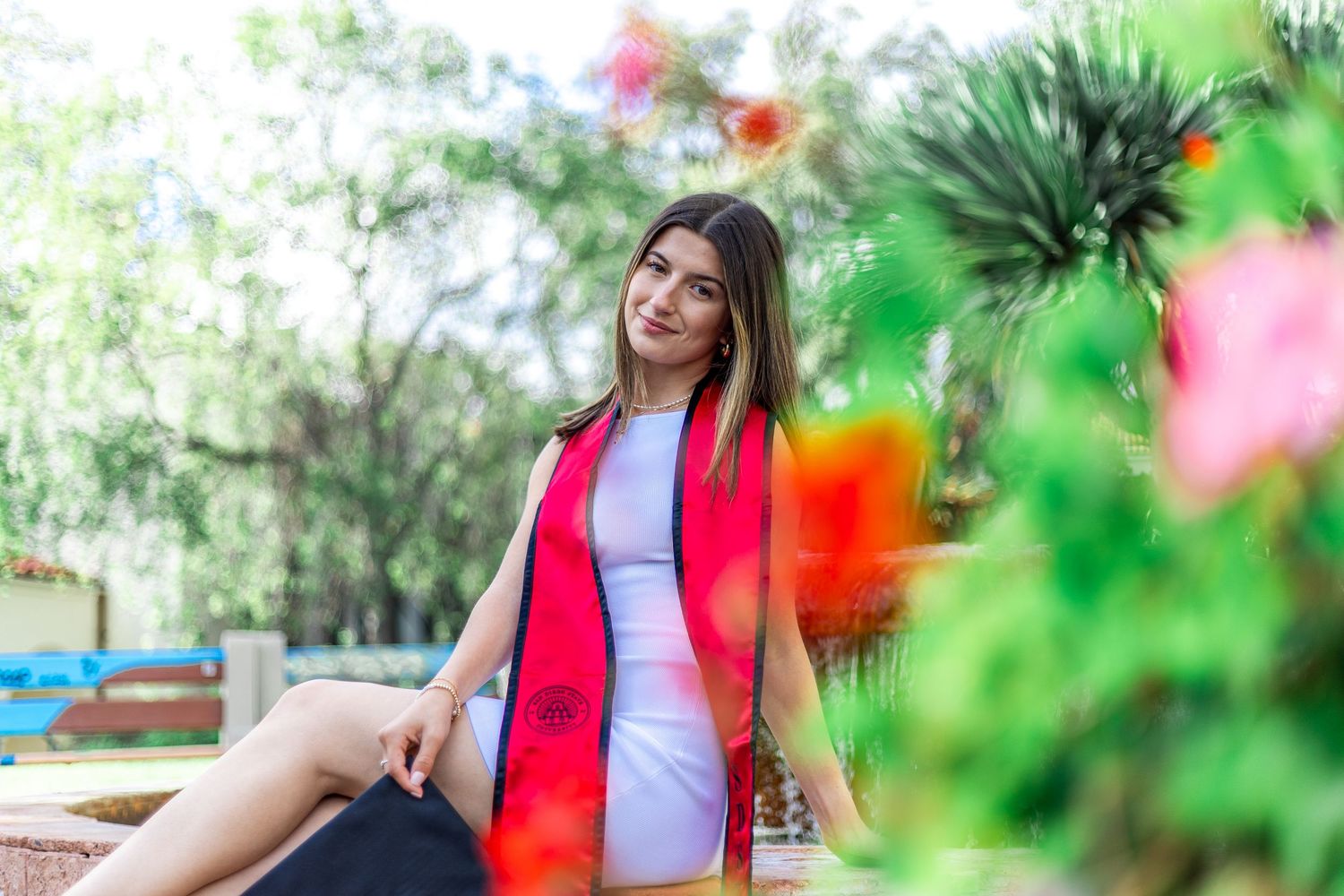 Graduate in white dress and red graduation stole poses joyfully in outdoor garden setting.