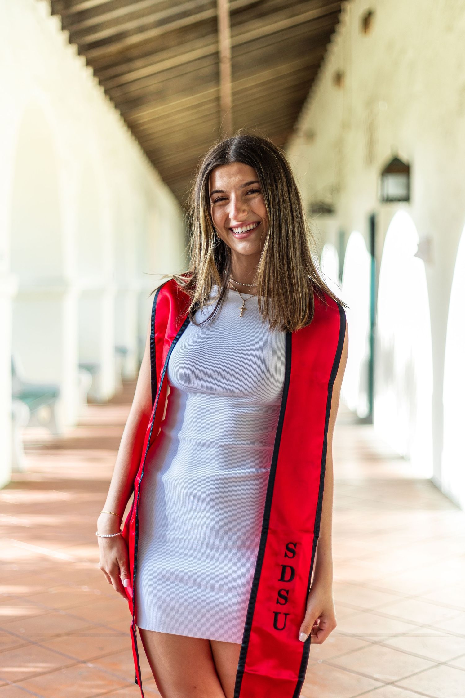 Graduate in white dress and red graduation stole poses in covered walkway.