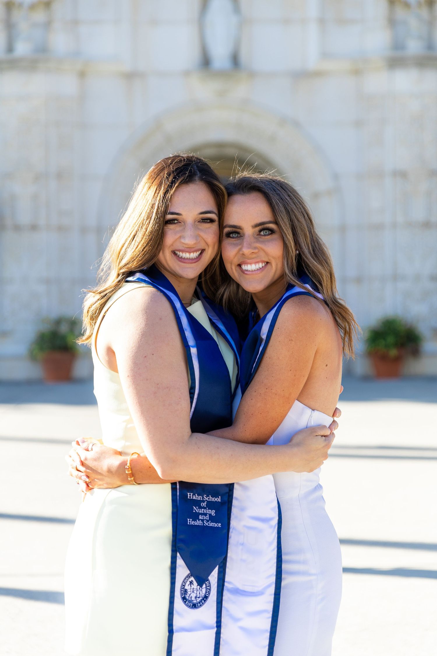 Two graduates in white dresses and blue stoles embrace while celebrating on sunny campus.