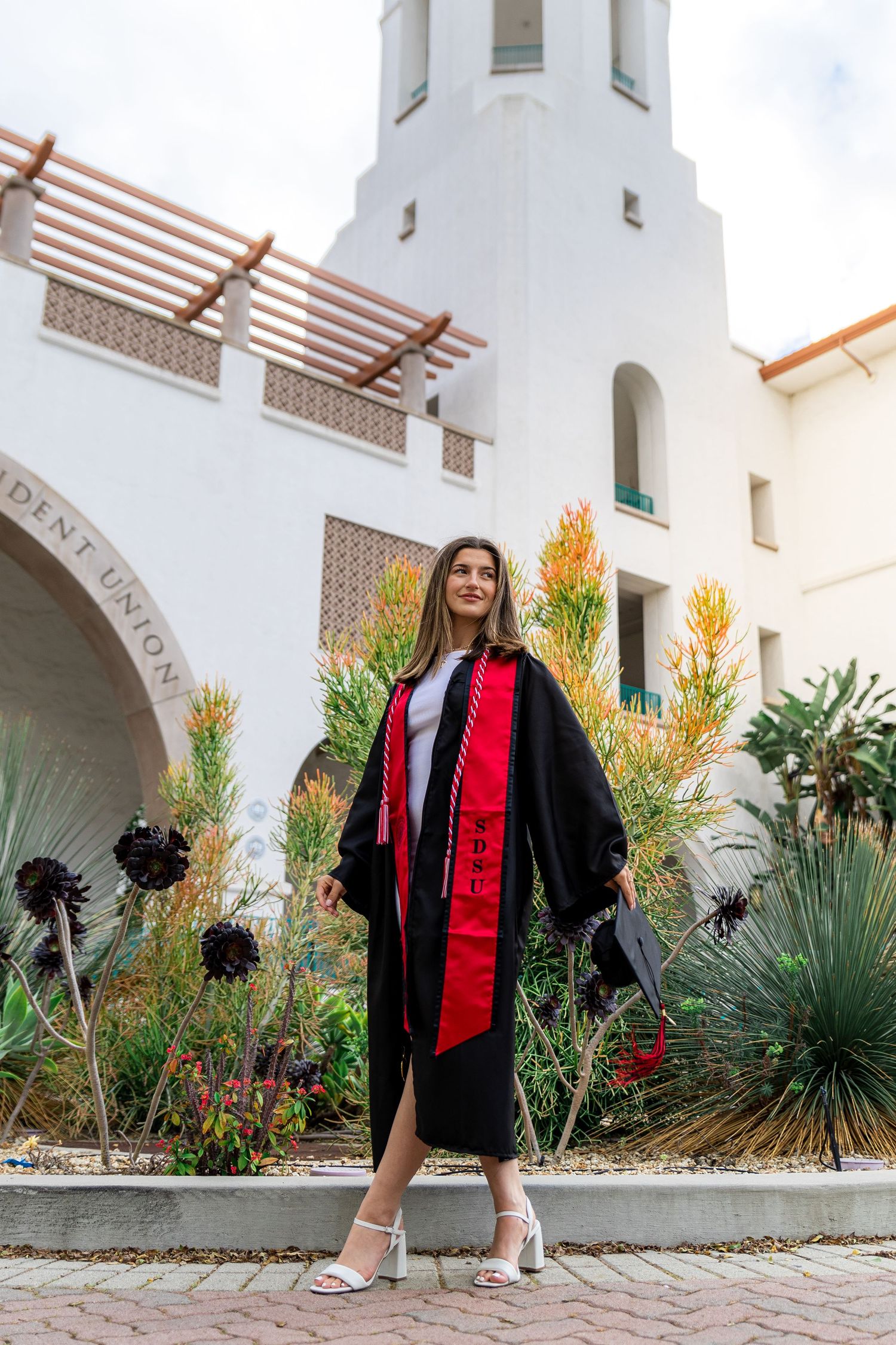 Graduate poses in front of Spanish-style architecture with desert landscaping.