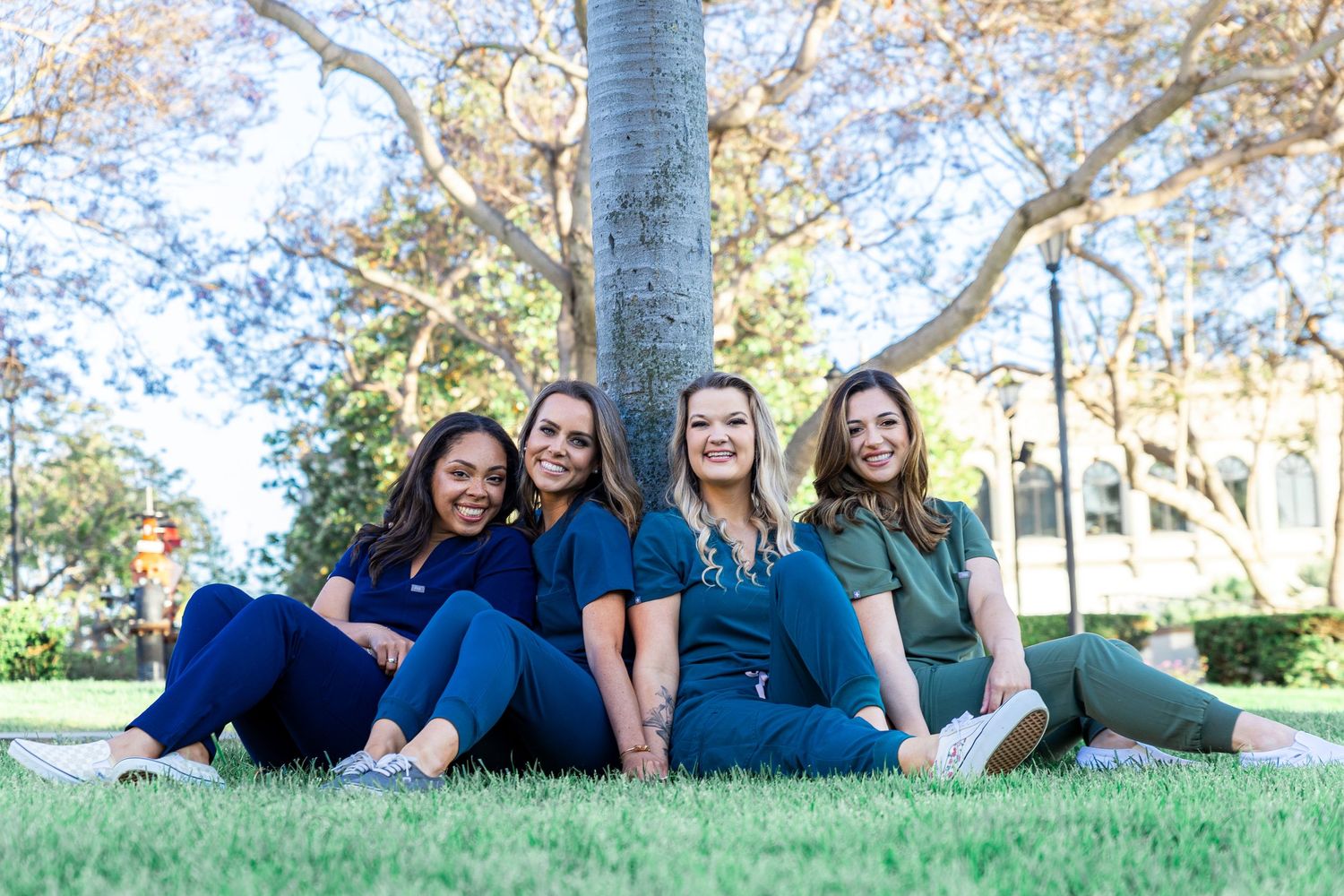 Group of graduates in blue scrubs sit together under tree on campus lawn.