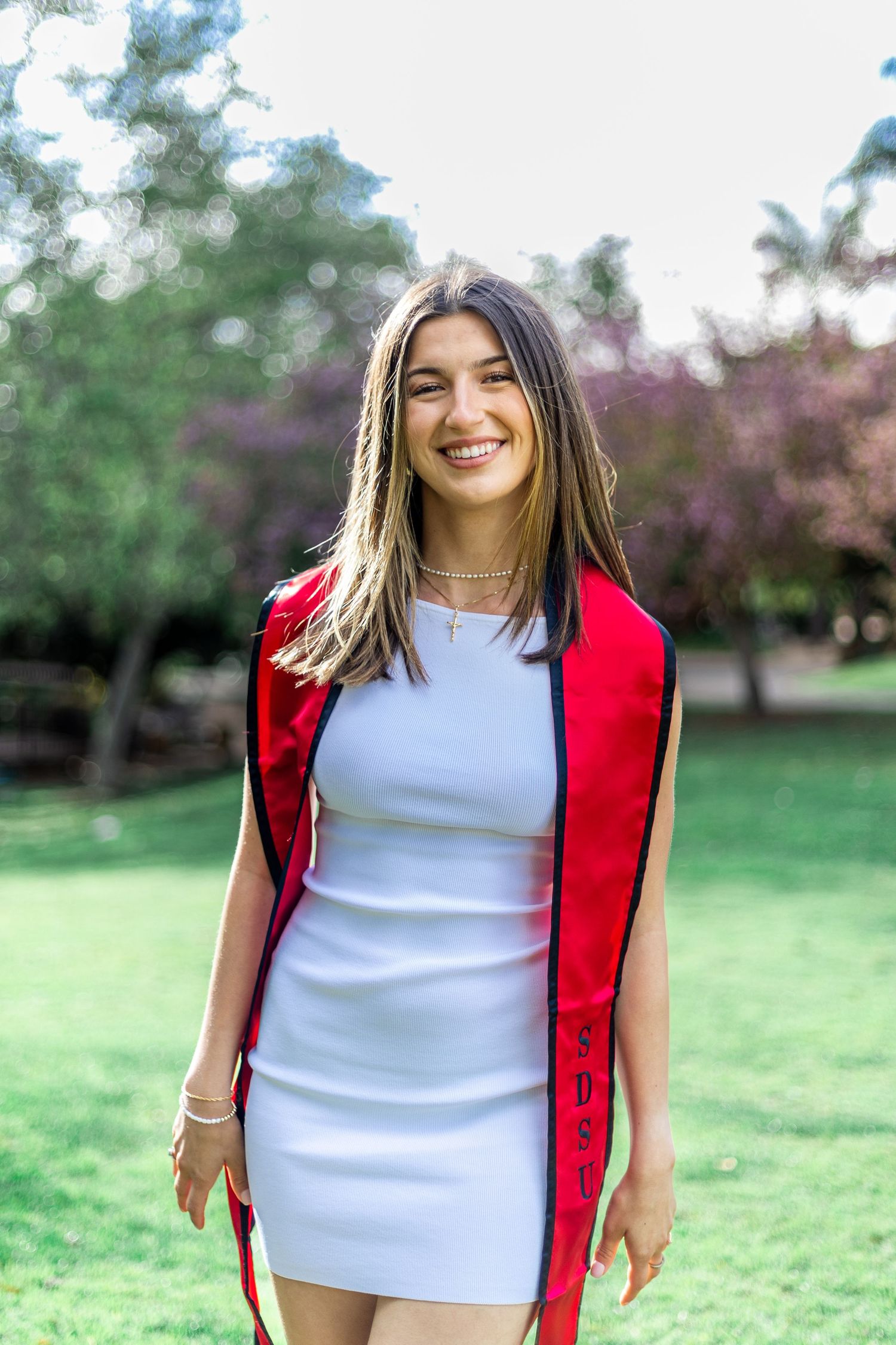 Graduate wearing red stole and white dress smiling outdoors in garden setting.