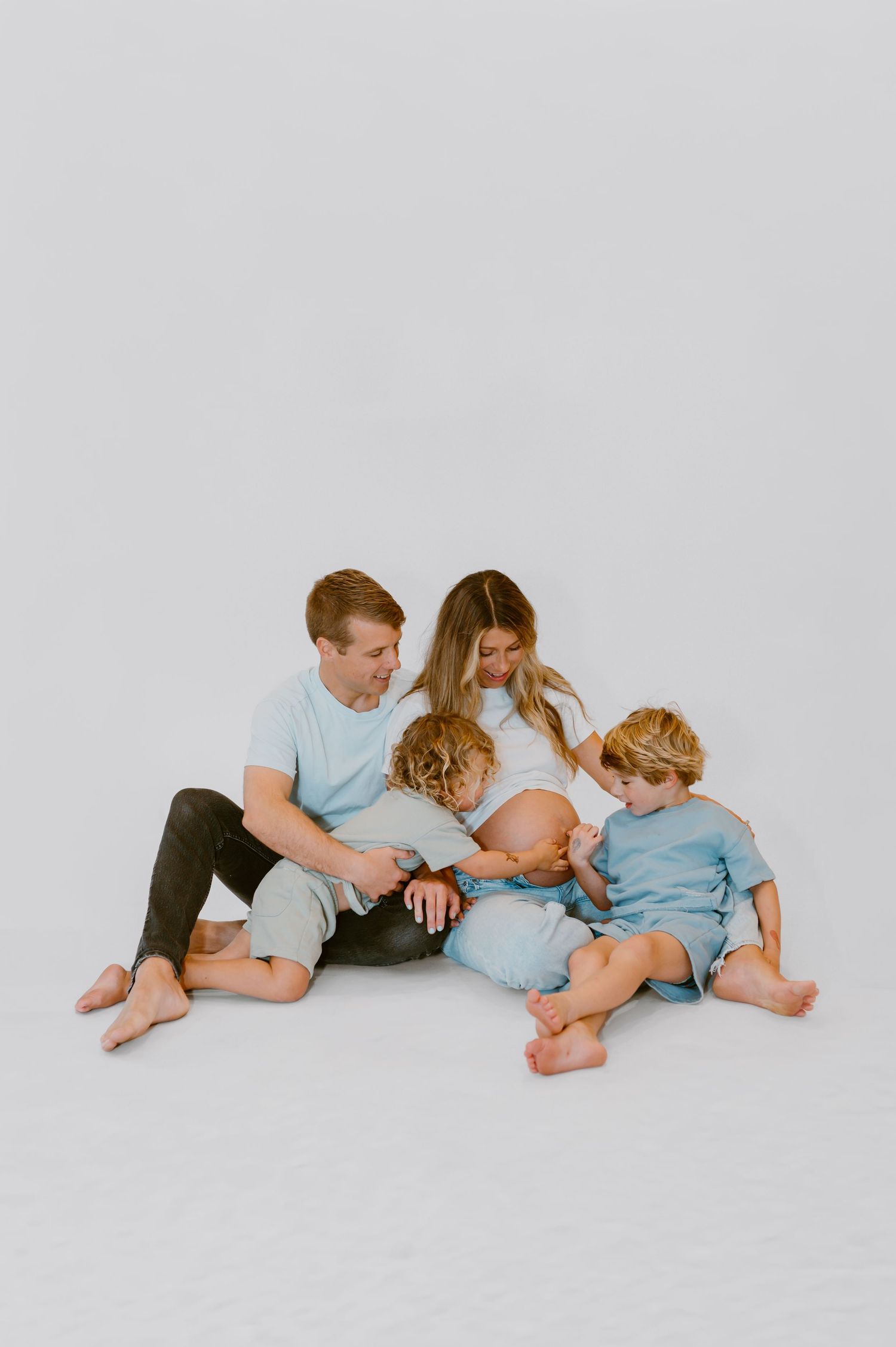 Family photo in studio in Yulee, Florida where toddler pokes mothers pregnant belly while laughing with his brother and dad