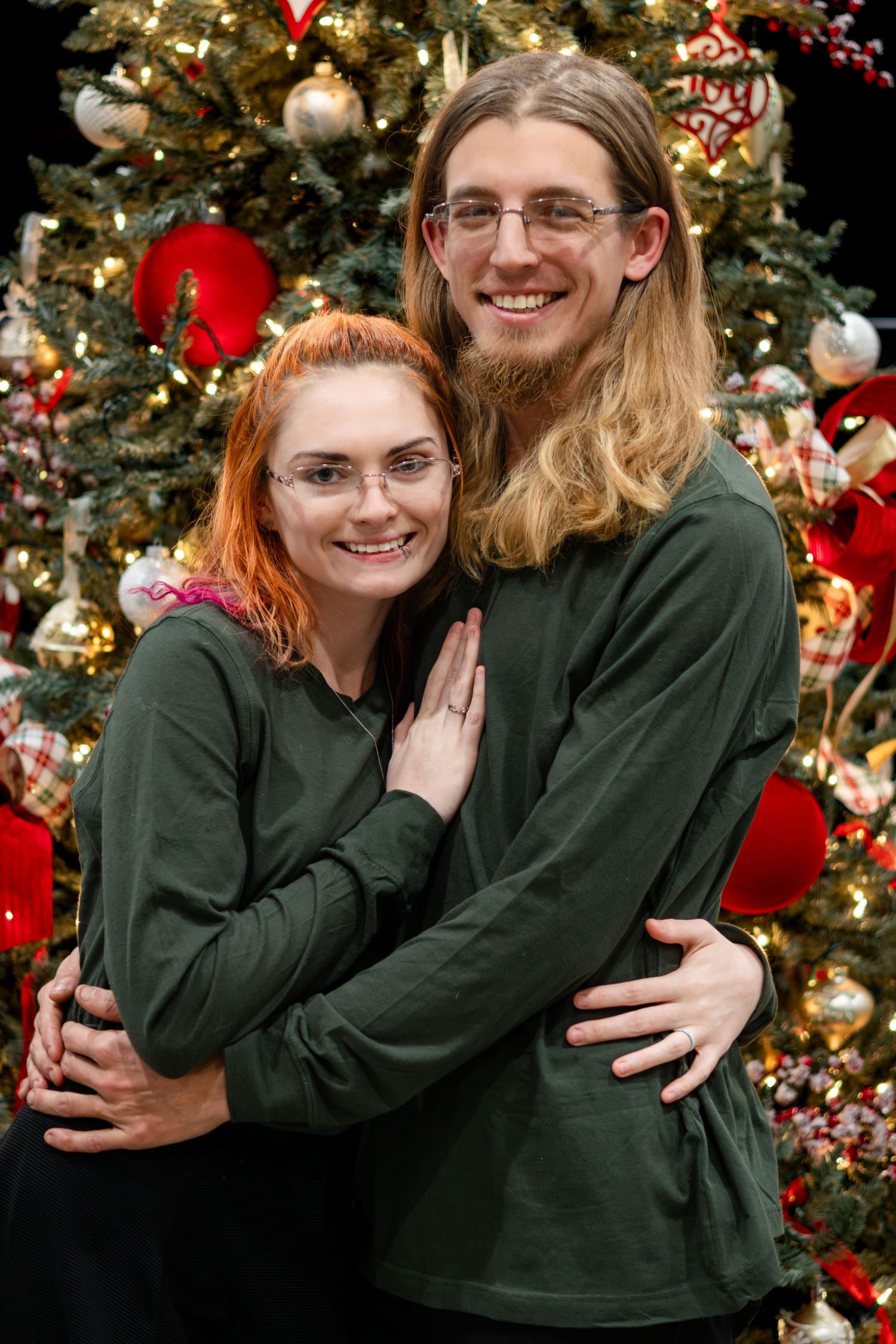 A couple in matching green sweaters embraces in front of a decorated Christmas tree with ornaments and lights.