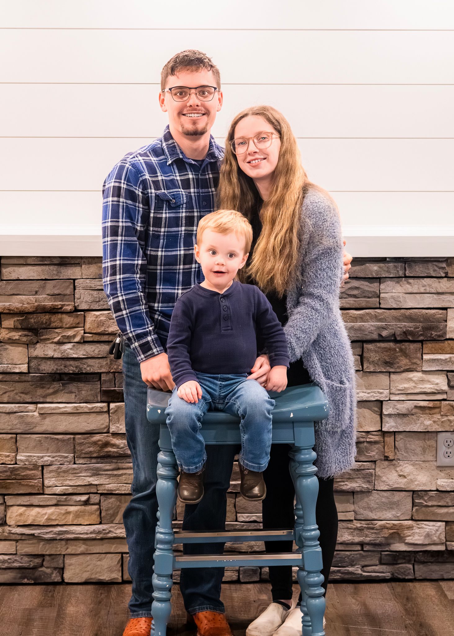 Small family posing together on blue stool against white shiplap and stone wall.