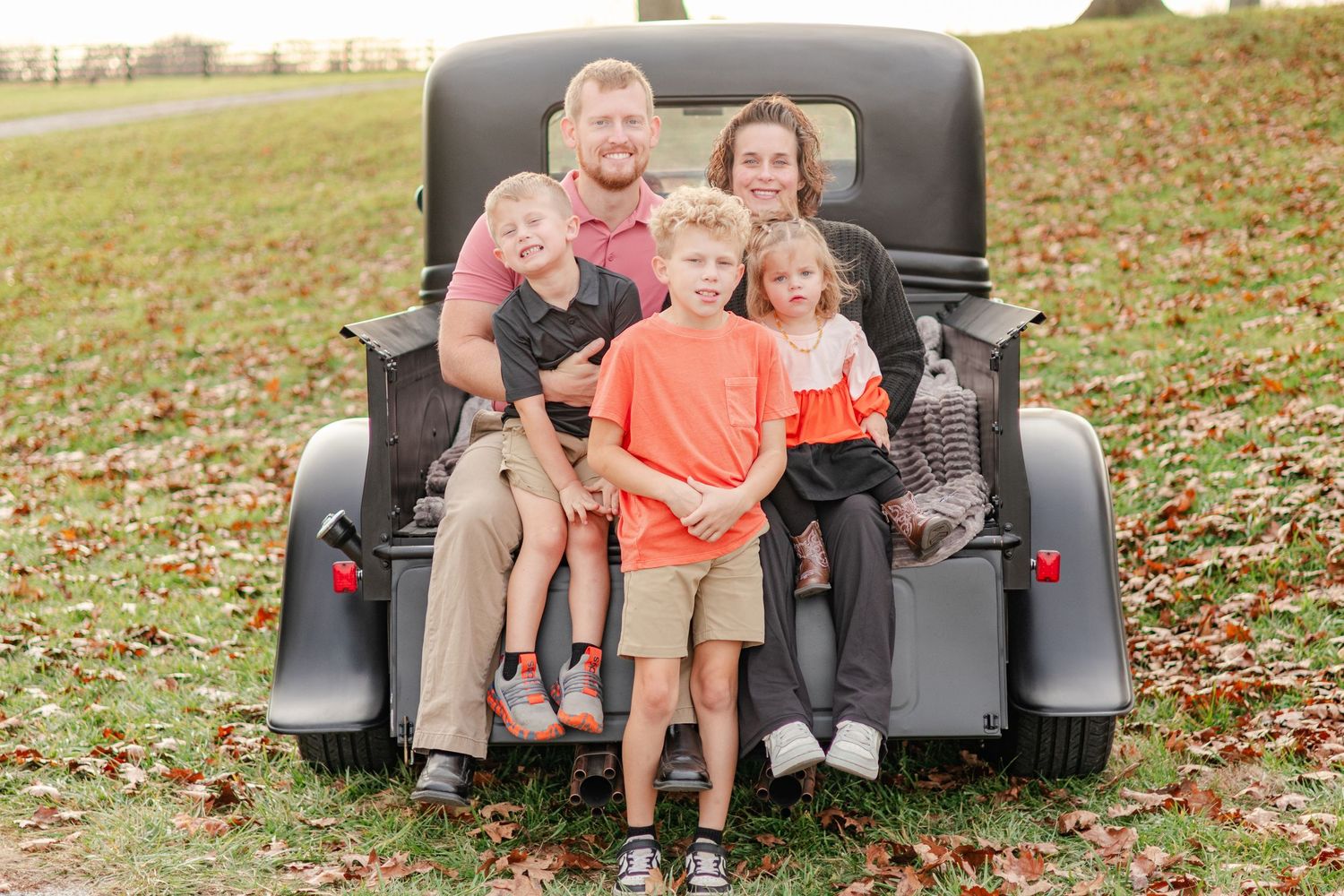 Family sitting together on a vintage pickup truck tailgate surrounded by fall leaves.