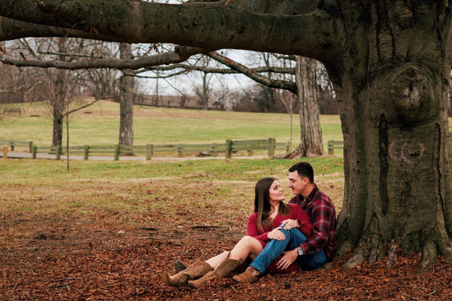 A couple sits together under autumn trees on fallen leaves in a park setting.