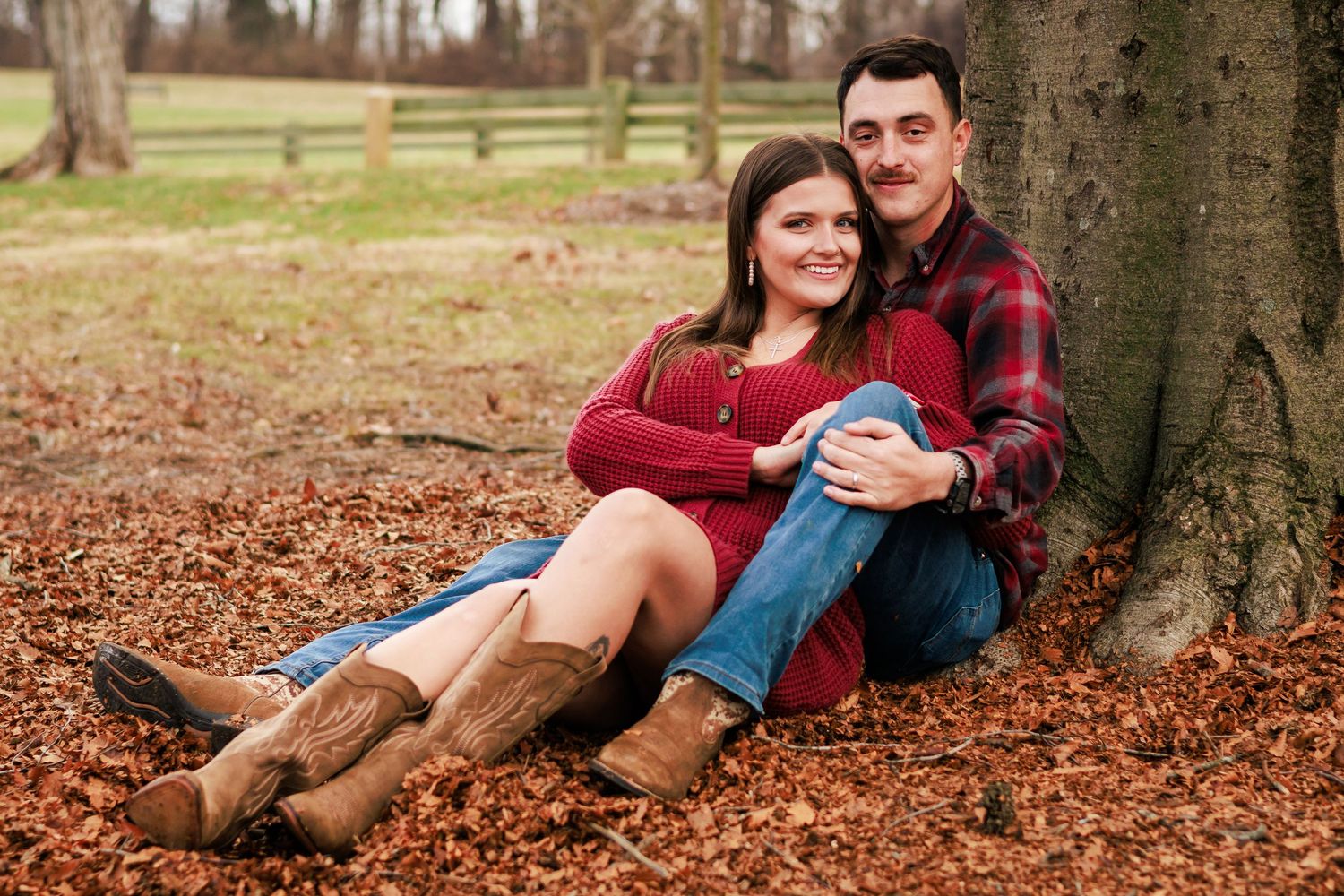 A couple relaxes on fallen autumn leaves near a large tree trunk in a park.