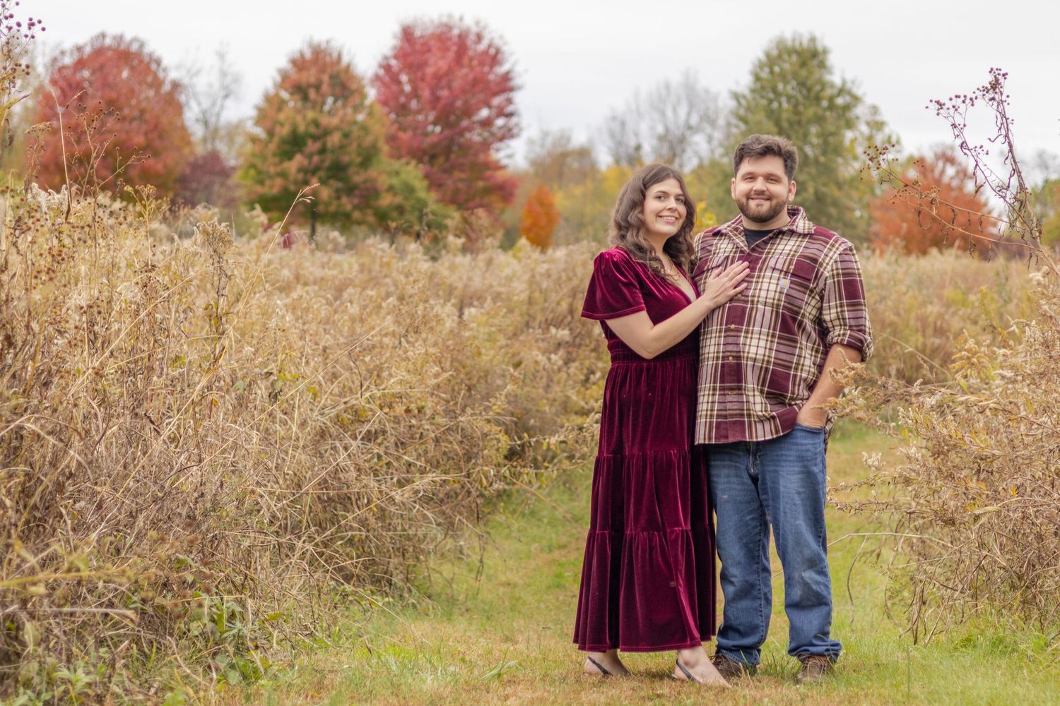 A couple poses together in a field during autumn with fall colors visible in the background.