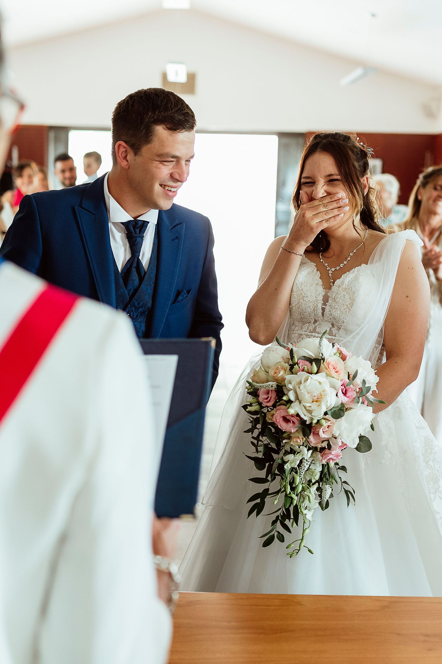 Les mariés rient joyeusement pendant leur cérémonie ; la mariée, vêtue d'une robe en dentelle blanche, tient un bouquet rose et blanc.