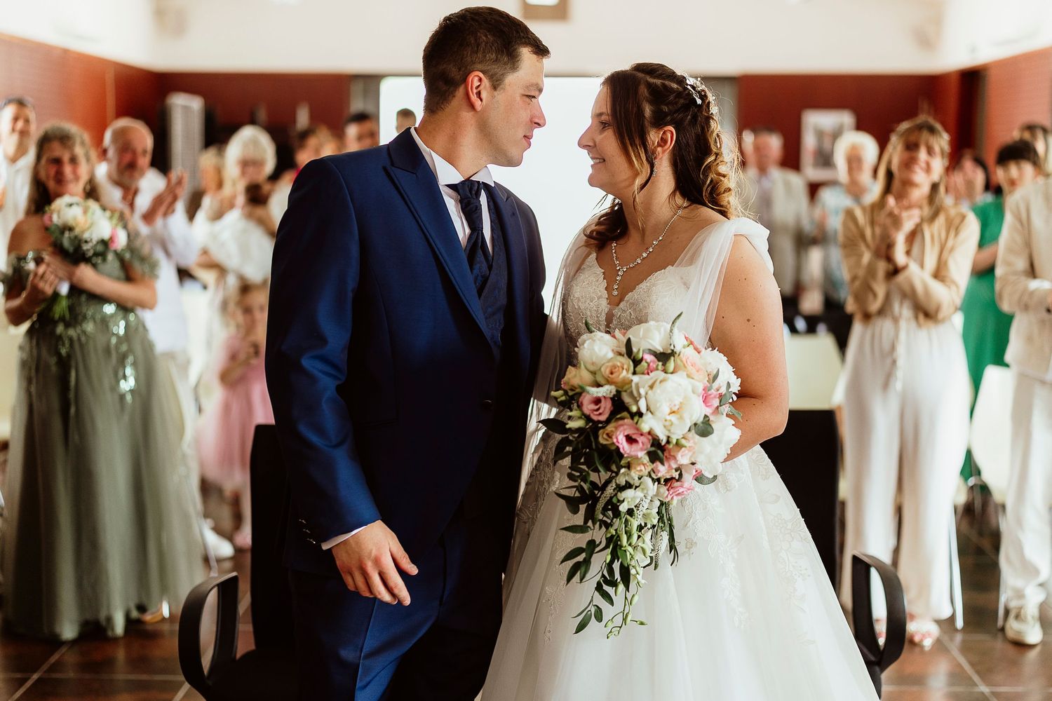 Un couple de jeunes mariés partage un moment romantique en remontant l'allée, entouré d'invités qui les applaudissent lors du mariage.