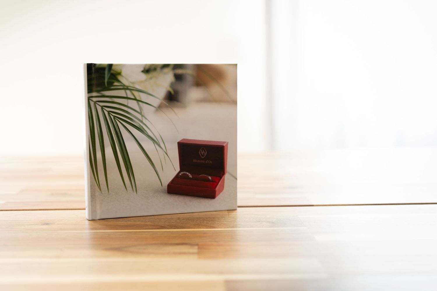 Un album photo carré posé sur une table en bois, avec une boîte à bijoux rouge contenant des alliances et une feuille de palmier verte.