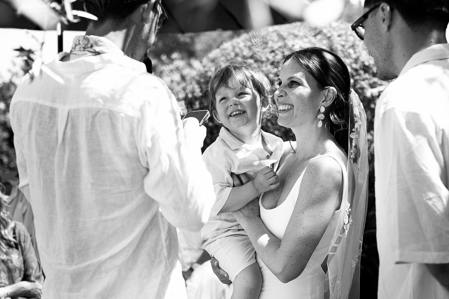 Photo en noir et blanc d'une mariée souriante en robe blanche, tenant un tout-petit qui rit lors d'une célébration de mariage en plein air.