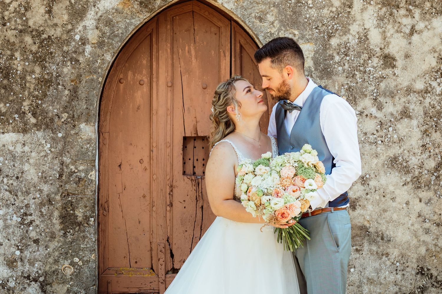 Les mariés partagent un moment romantique devant une porte en bois rustique surmontée d'une arche ; la mariée tient un bouquet aux tons pêch