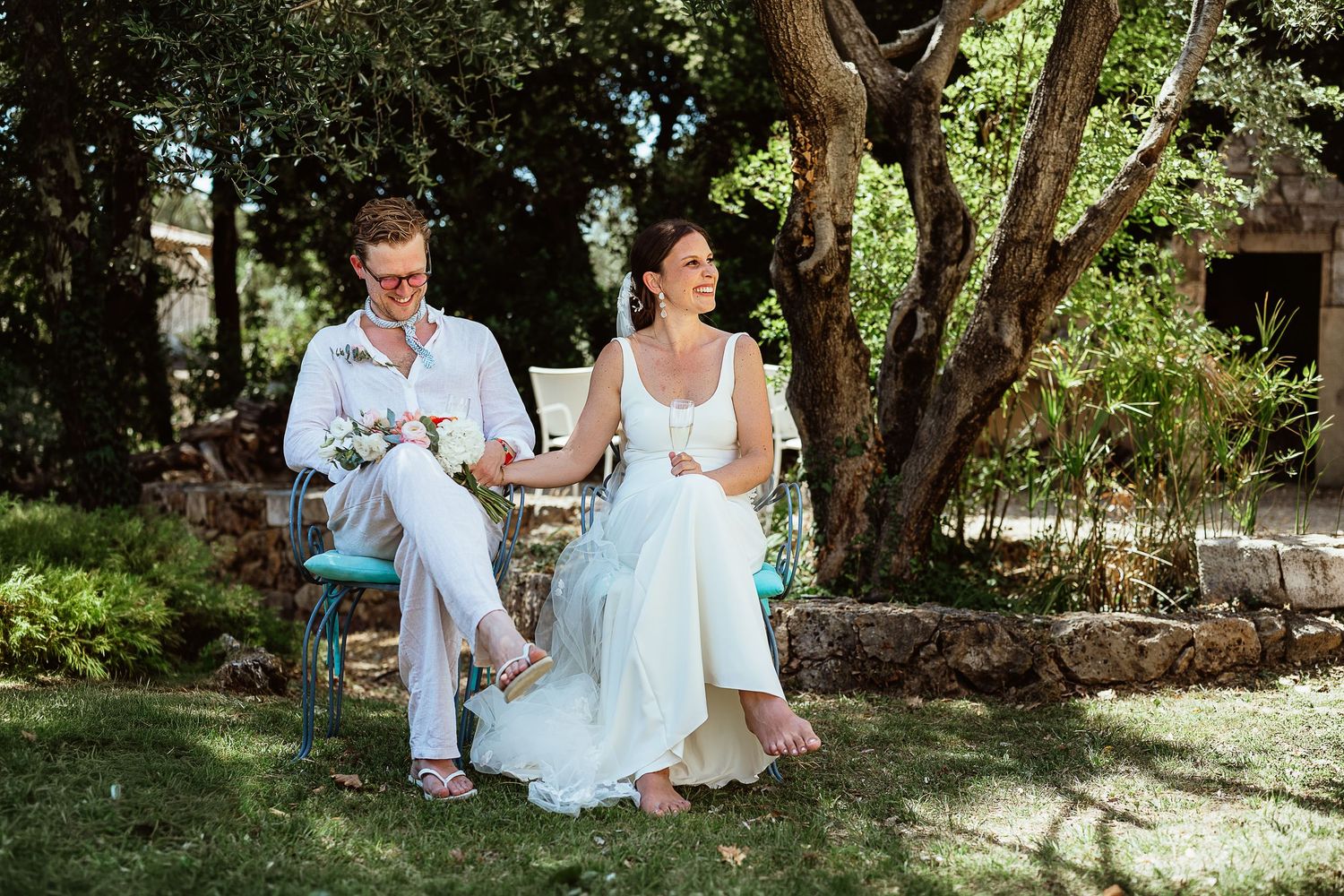 Les mariés sont assis sur des chaises dans un jardin, lors d'une cérémonie de mariage en plein air, entourés d'arbres au feuillage verdoyant