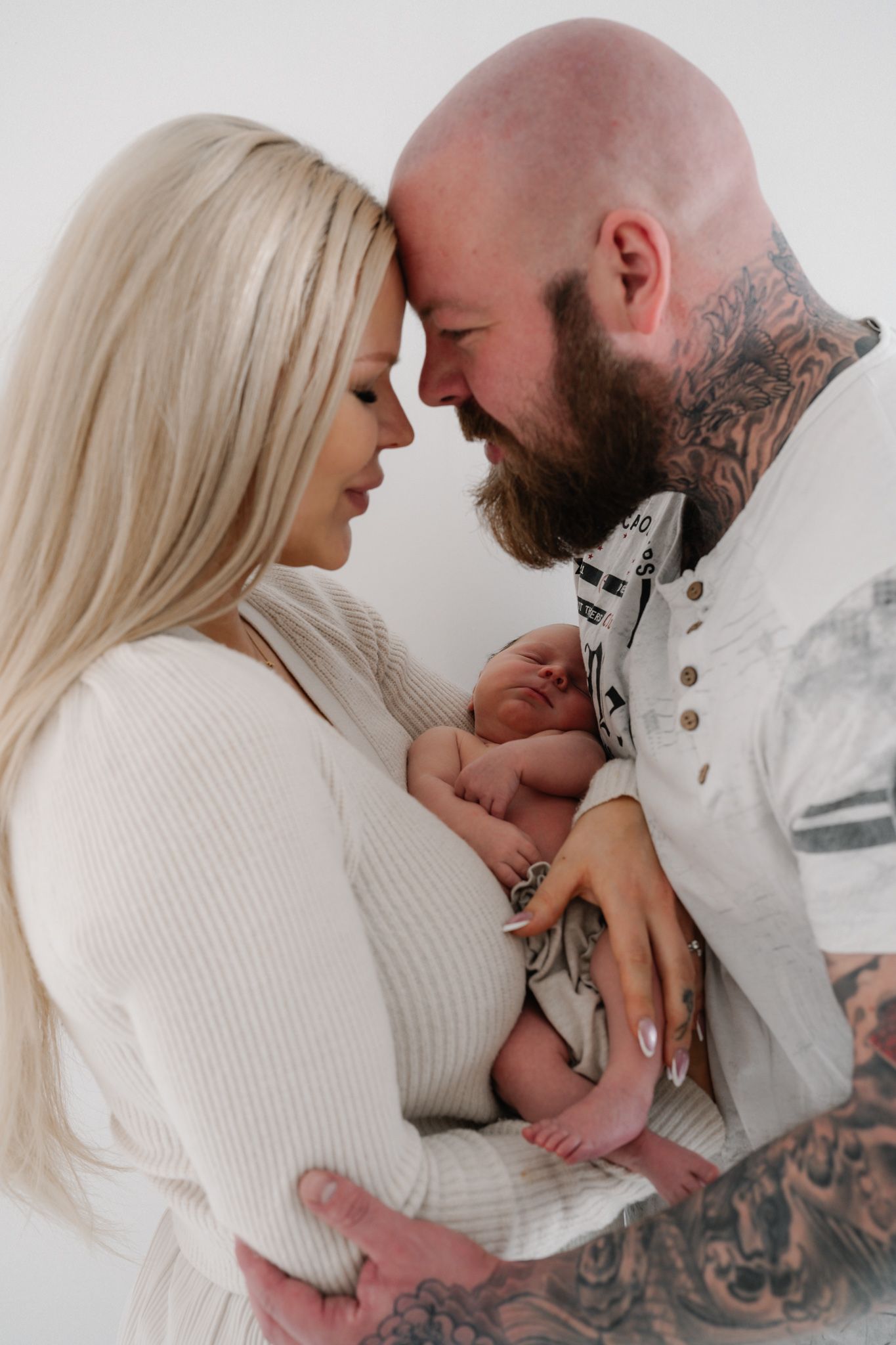A tattooed couple with long blonde hair tenderly holds their newborn baby, foreheads touching in a white studio.