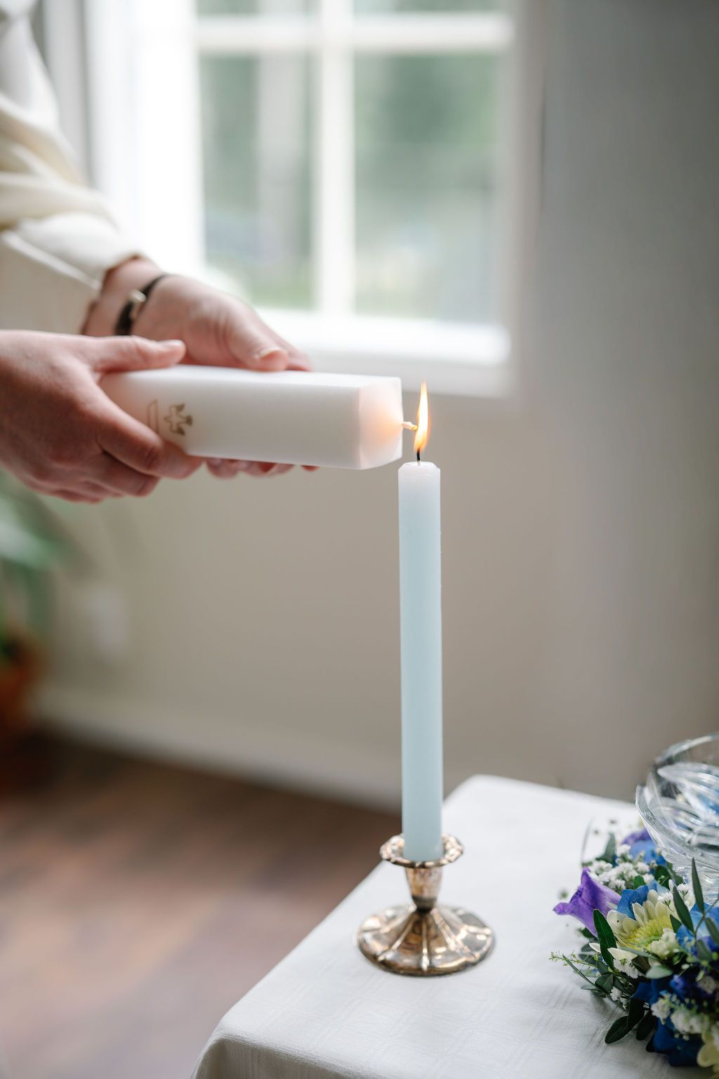 Hands lighting a white taper candle in a gold candlestick holder on a white table with flowers nearby.