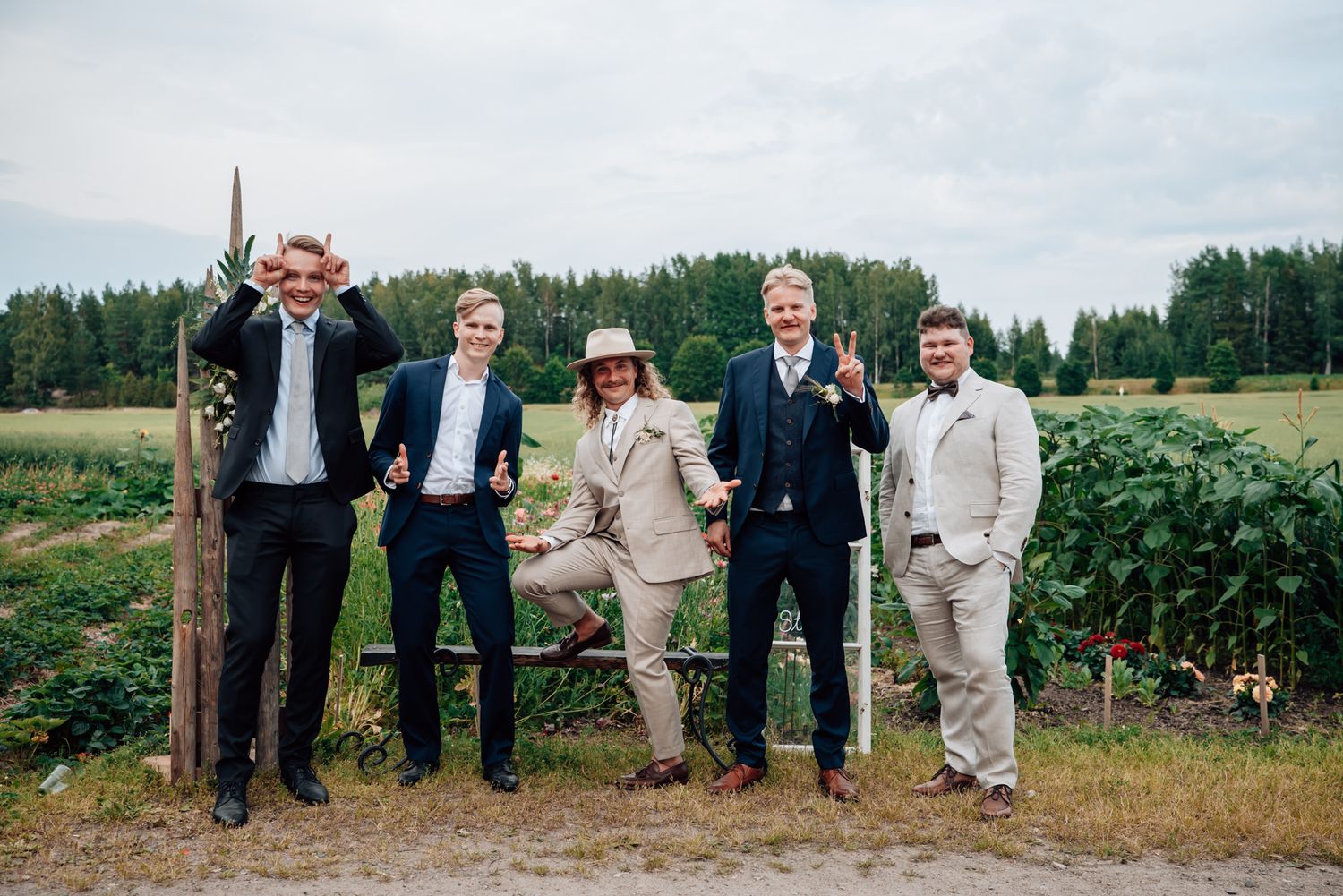 Five men in formal suits posing playfully outdoors at a wedding celebration with green fields in the background.