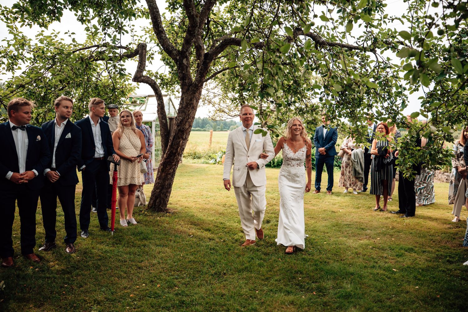 Bride and groom walk together outdoors at a garden wedding ceremony surrounded by guests under apple trees.