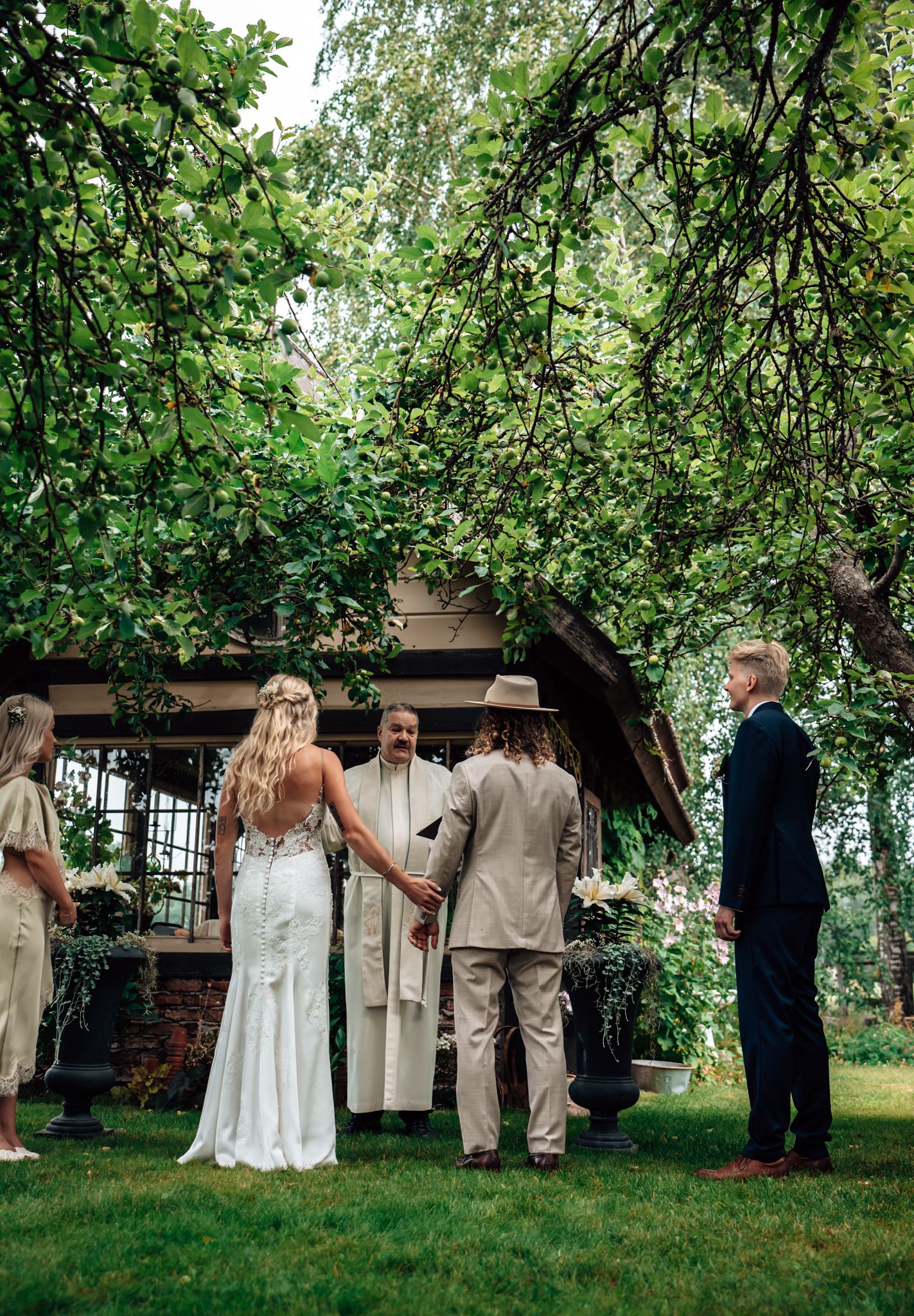 Outdoor garden wedding ceremony with bride in white gown, groom in tan suit, and officiant under lush green trees.