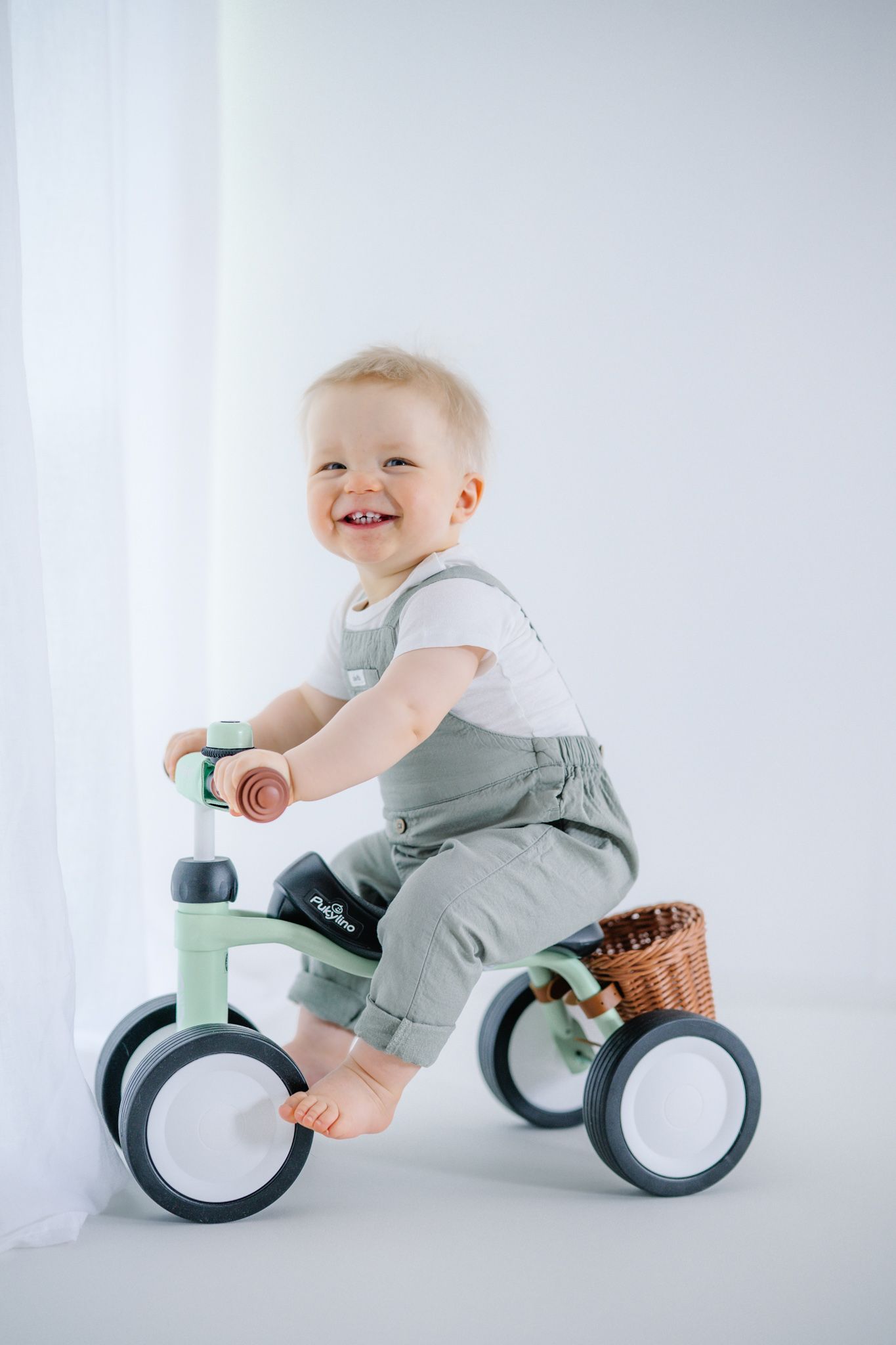 Smiling baby in green overalls riding a mint-colored balance bike with a wicker basket in a white room.
