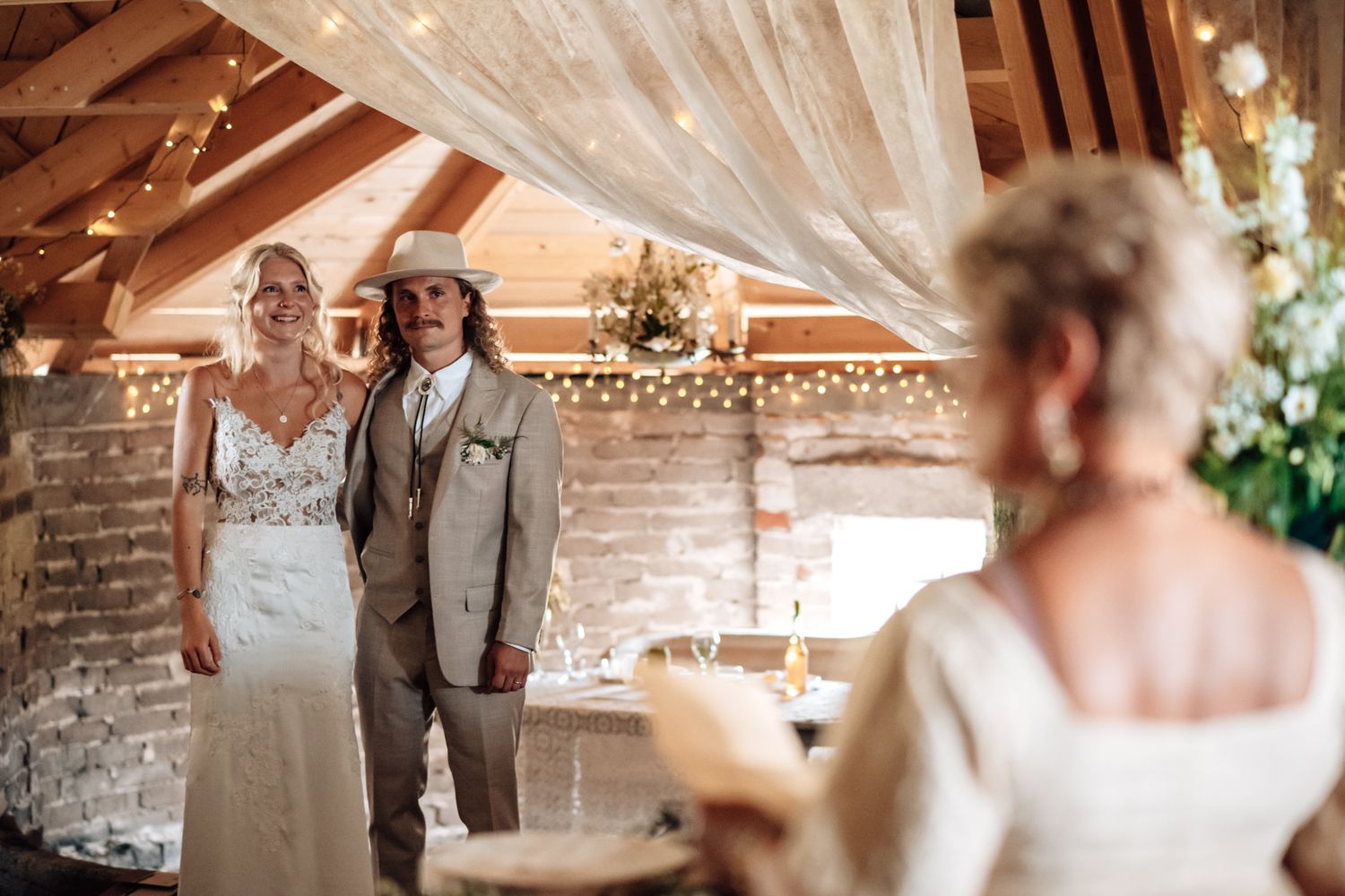 Bride and groom smiling during rustic barn wedding ceremony with fairy lights and exposed brick backdrop.