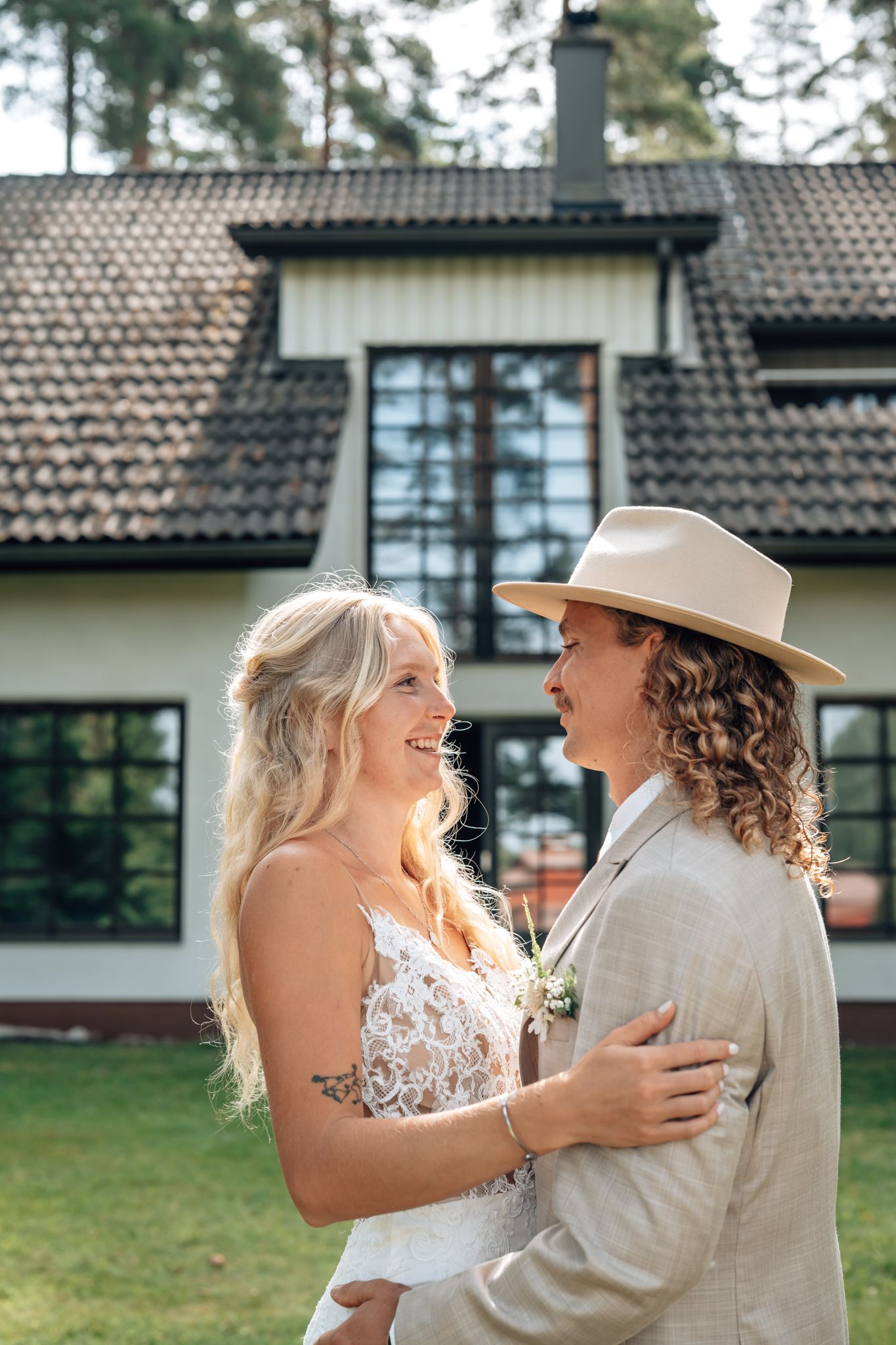 A smiling bride and groom in boho attire embrace outdoors in front of a charming house on their wedding day.